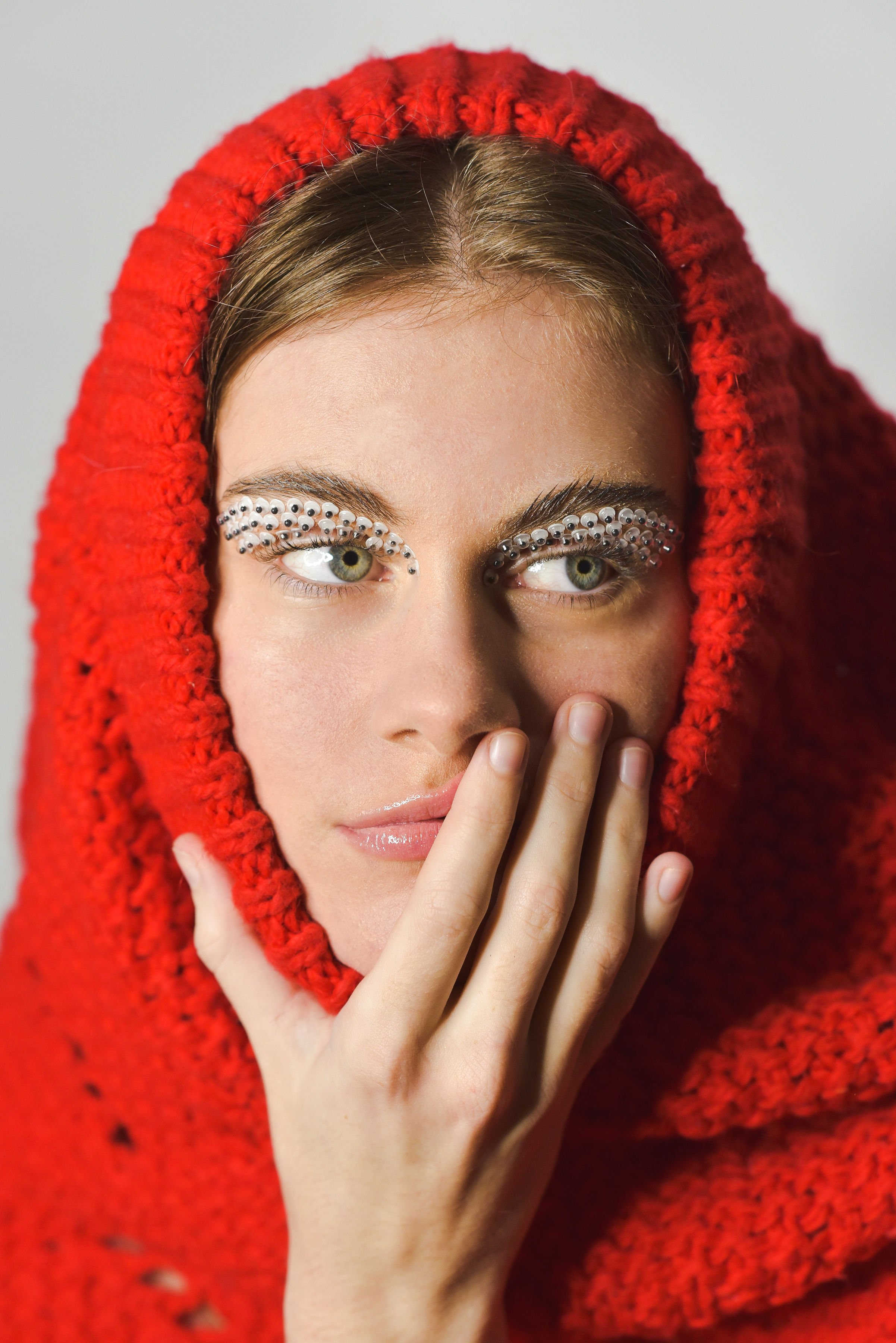 A woman wearing a red knit hooded scarf with rhinestones on her eyelids, touching her lips with her fingers.