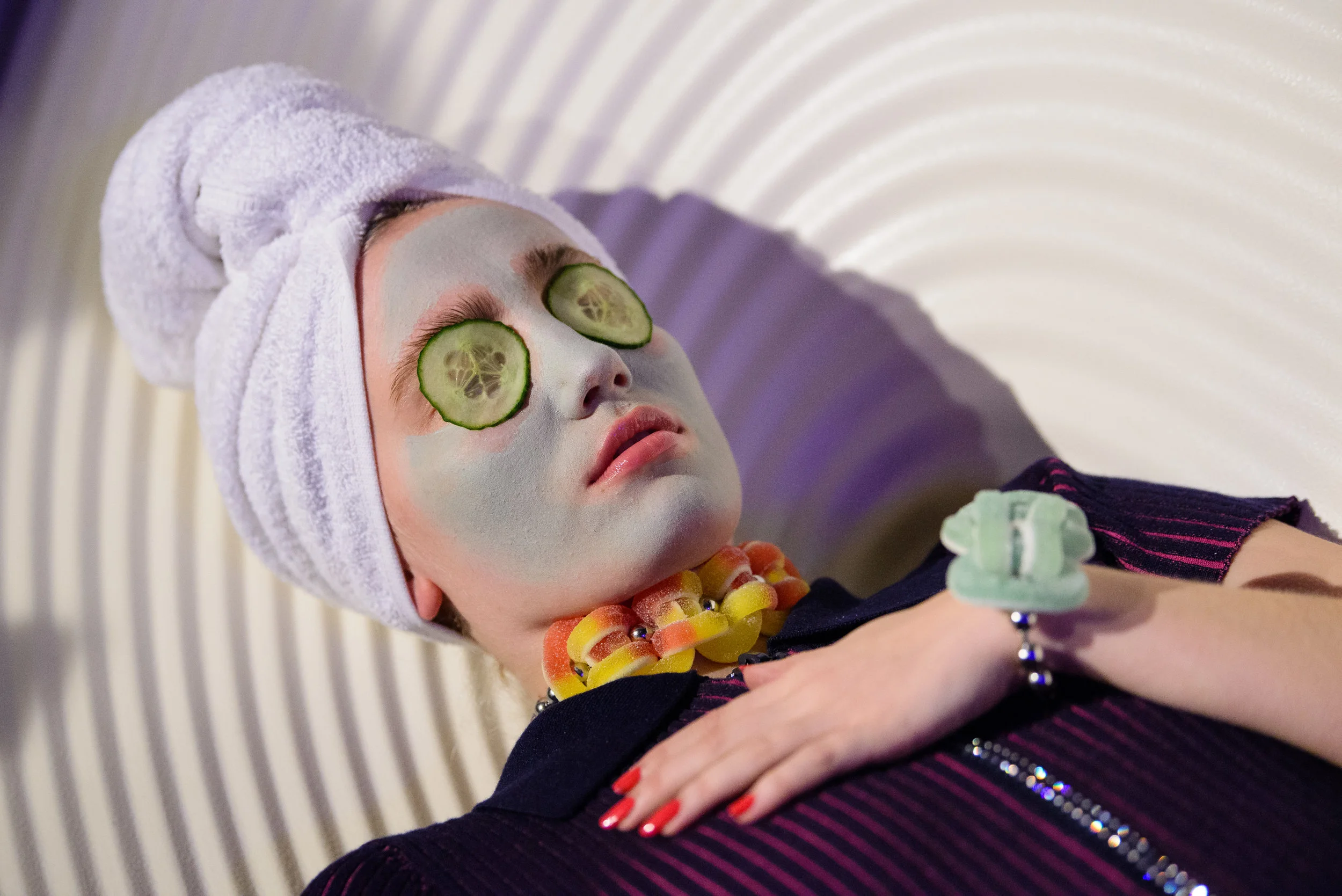 Young woman with face mask, cucumber slices over eyes, towel wrapped around head, colorful jewelry, lying on a textured surface with eyes closed.
