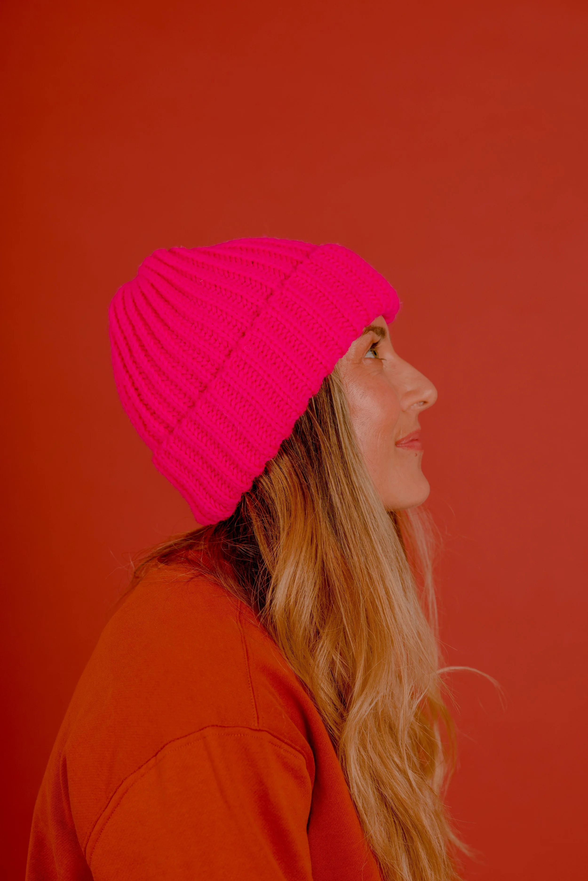 Side profile of a woman with long, wavy hair wearing a bright pink knit beanie and an orange sweatshirt, against a warm orange background.