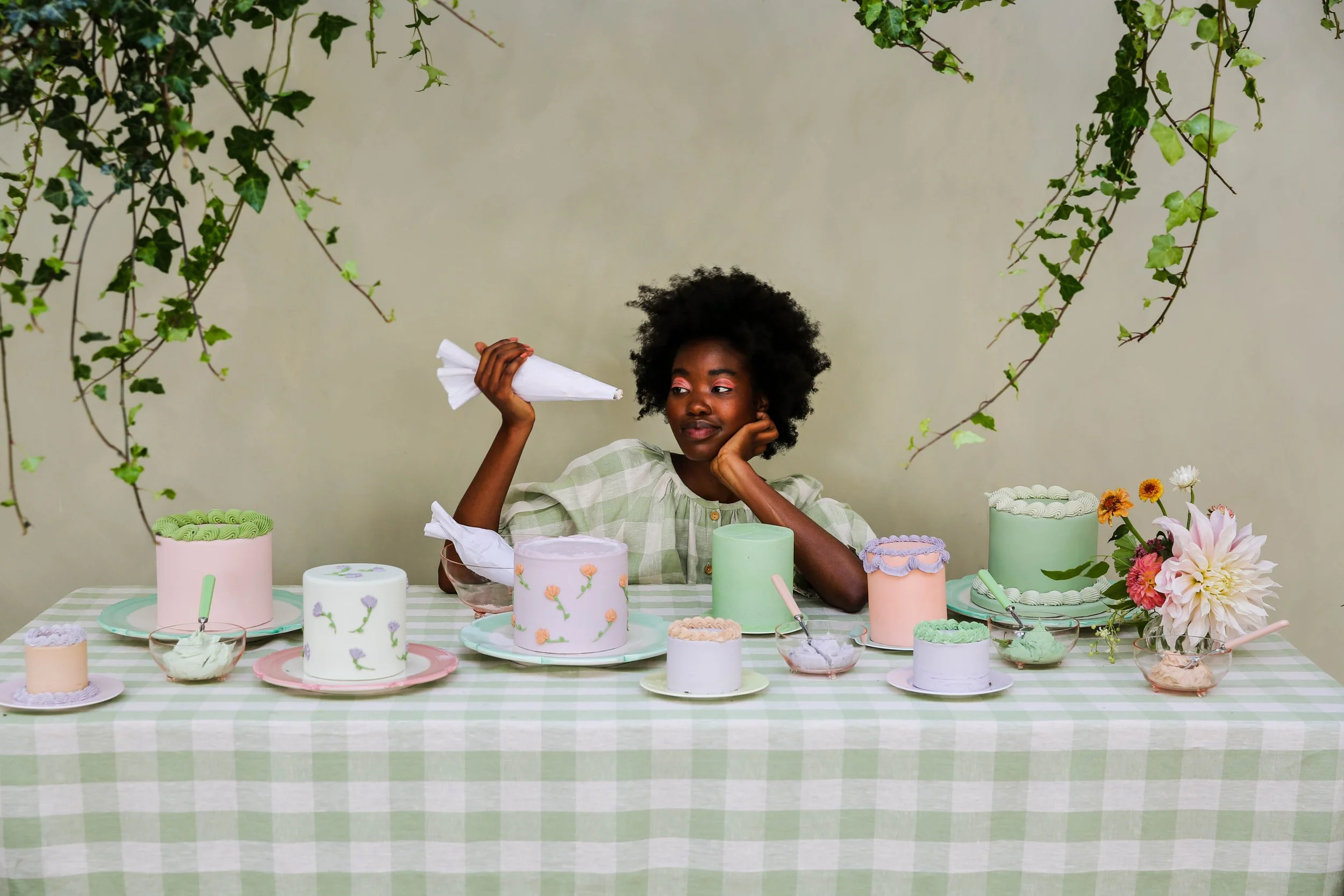 A woman with natural curly hair sits at a table with pastel-colored cakes, frosting, and flowers, holding a piping bag, surrounded by greenery.