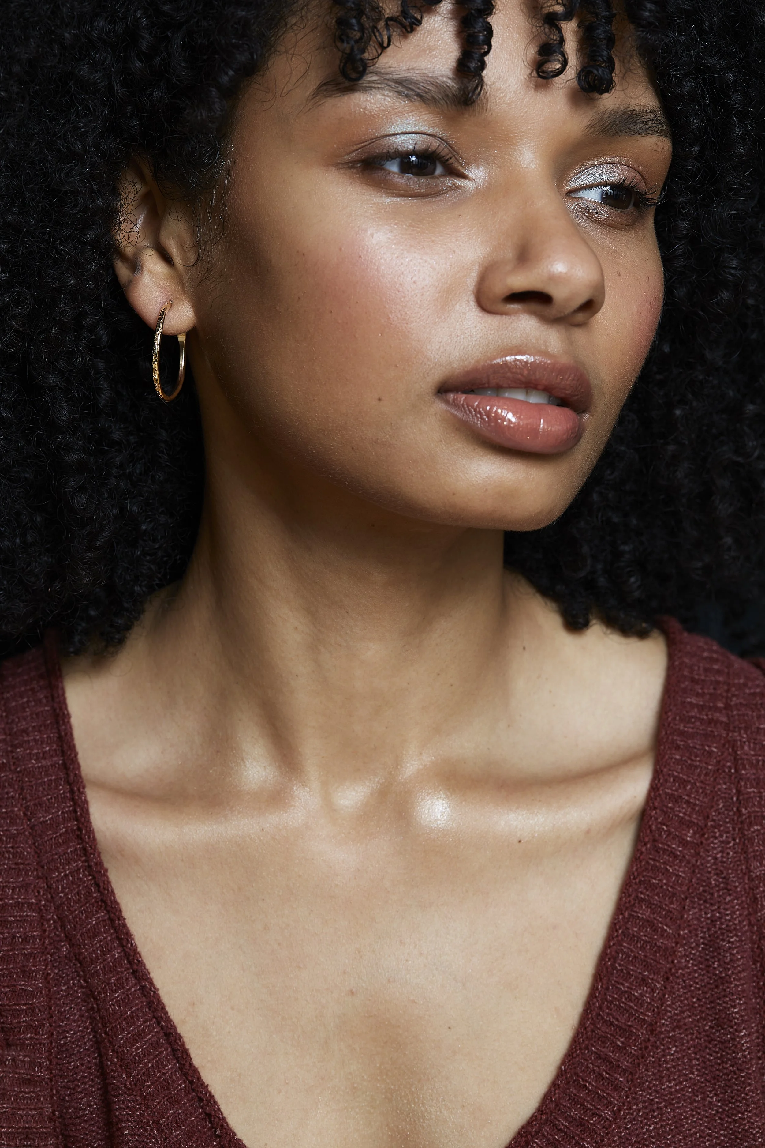 A young woman with dark, curly hair wearing hoop earrings and a maroon top, looking slightly to the side with a neutral expression.