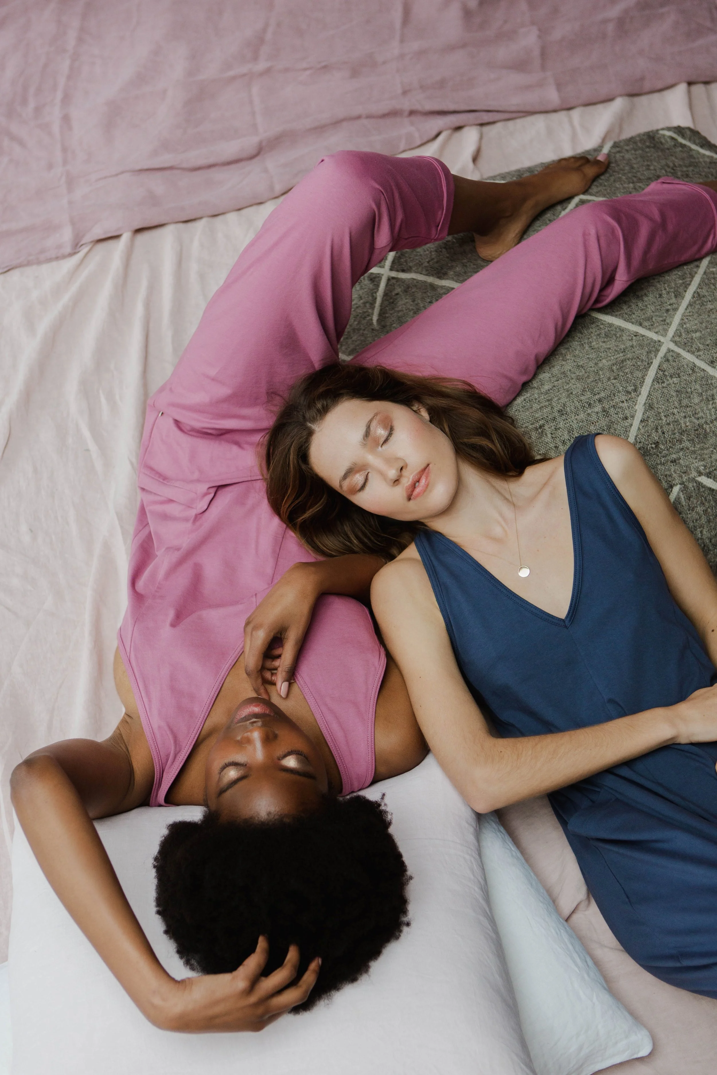 Two women lying on the floor together, their eyes closed, appearing relaxed and peaceful. One woman has curly black hair and is wearing a pink sleeveless top, while the other has straight brown hair and is wearing a blue sleeveless dress. They are re