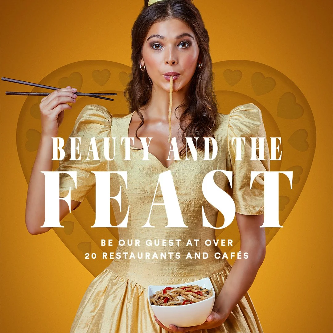 Woman in a gold dress holding a bowl of noodles with chopsticks, licking her lips against a yellow background with a heart graphic and promotional text for a restaurant guide.