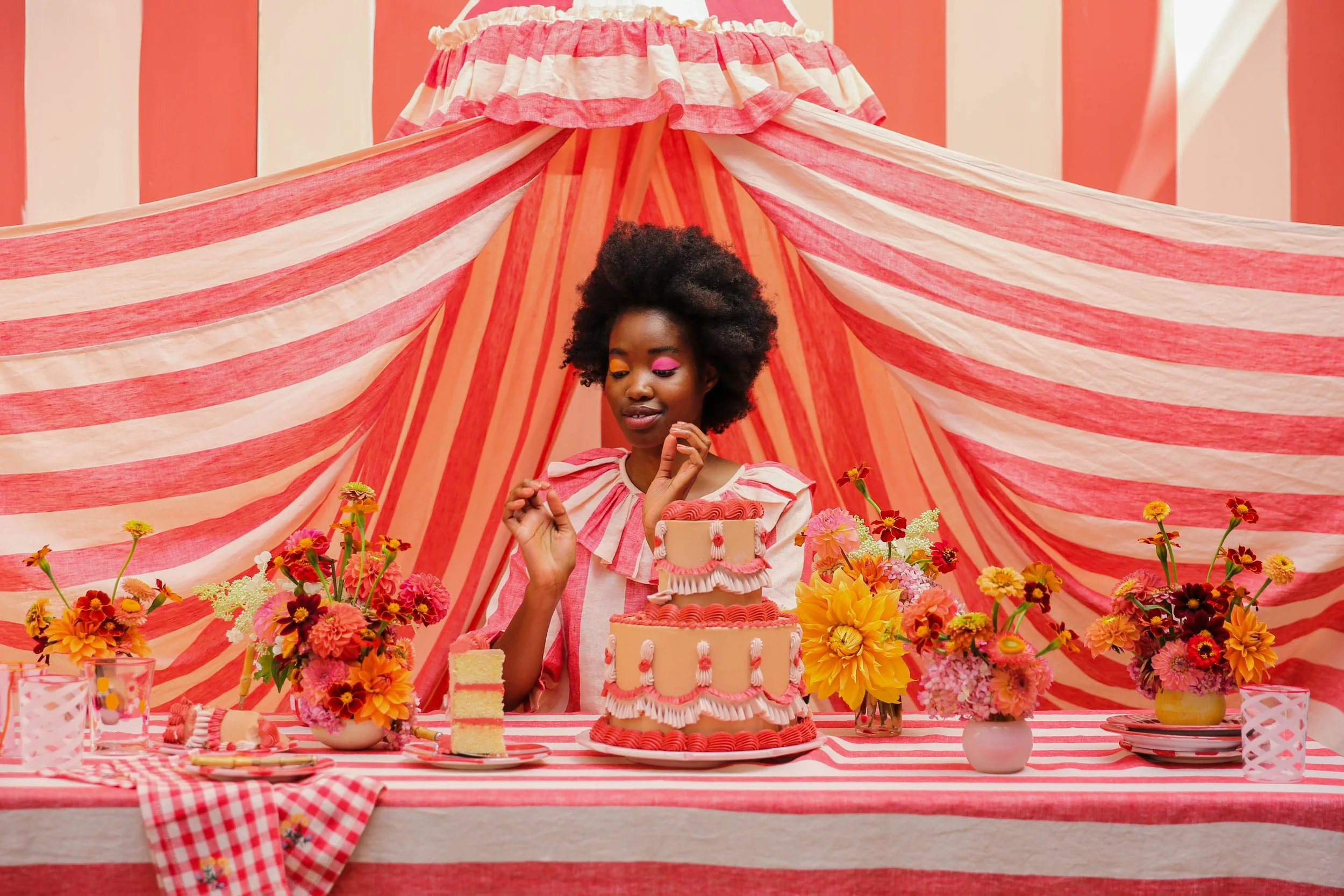A woman with an afro hairstyle is sitting at a table decorated with pink, red, and yellow flowers, with a pink and red striped cake in front of her, surrounded by pink and red floral arrangements and pink-striped tablecloths and backdrop.