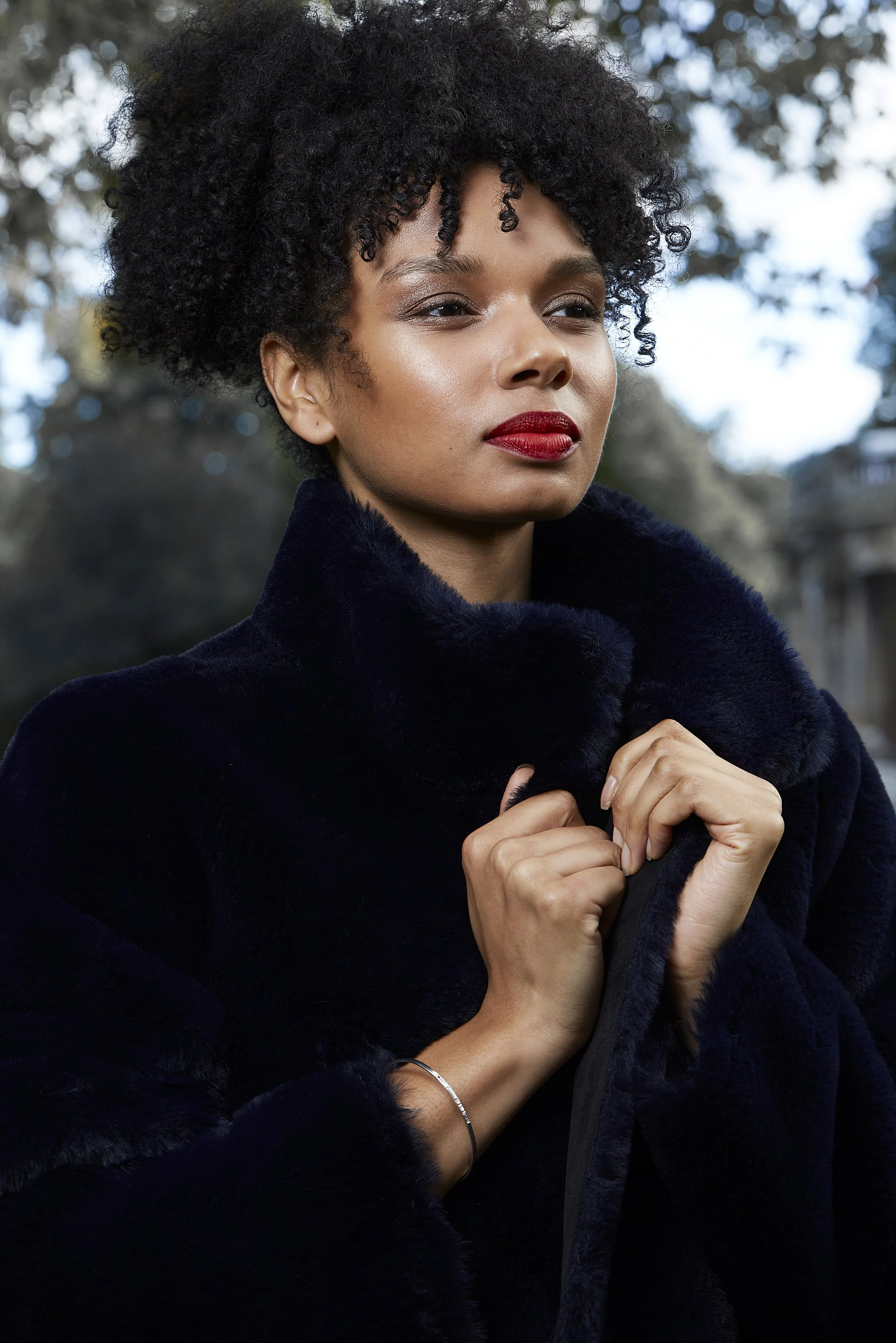 A woman with short curly black hair and red lipstick, wearing a dark fur coat, stands outdoors with trees in the background.
