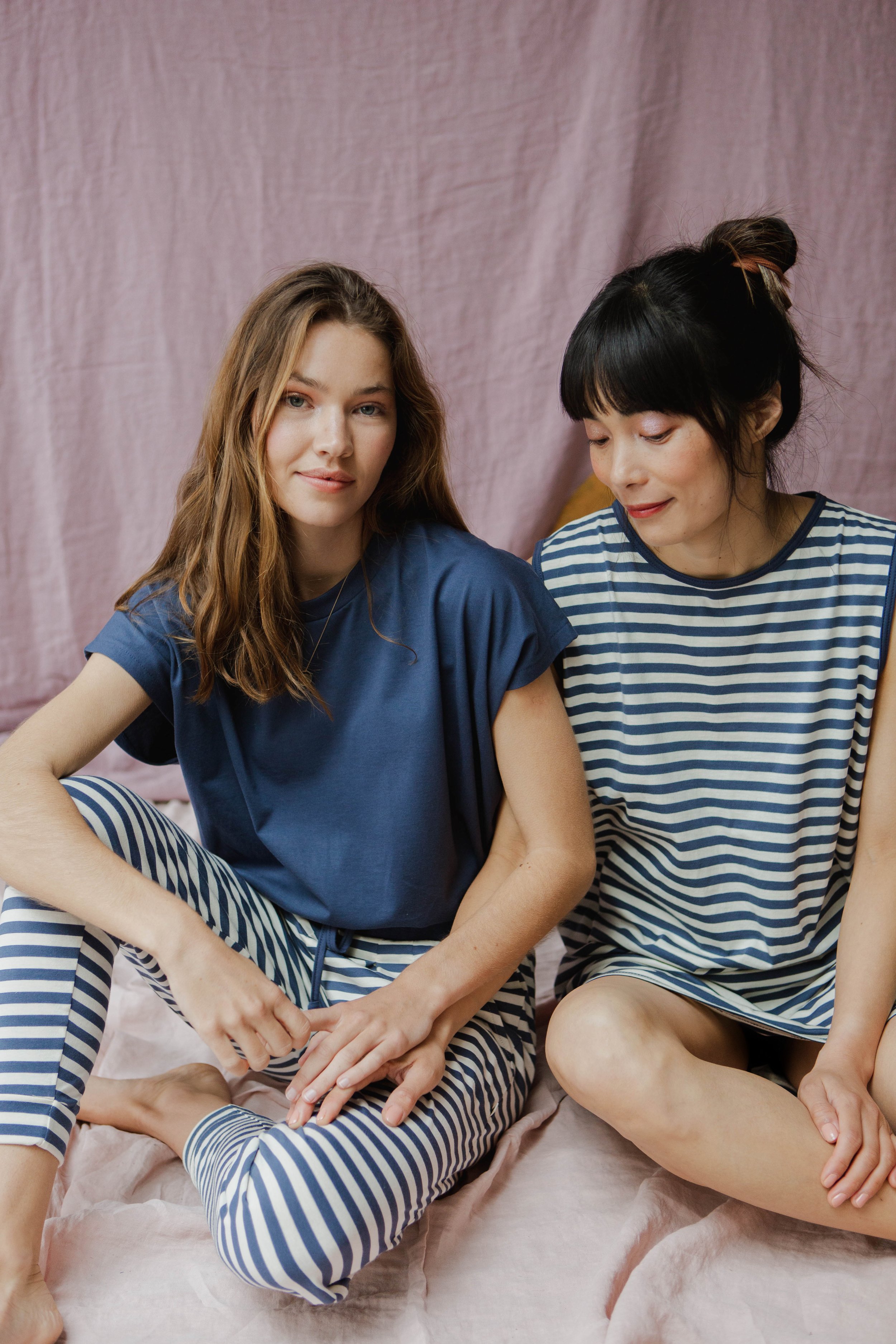 Two women sitting on a pink fabric backdrop, wearing blue and white striped pajamas, with one woman in a plain blue shirt, posing for the photo.