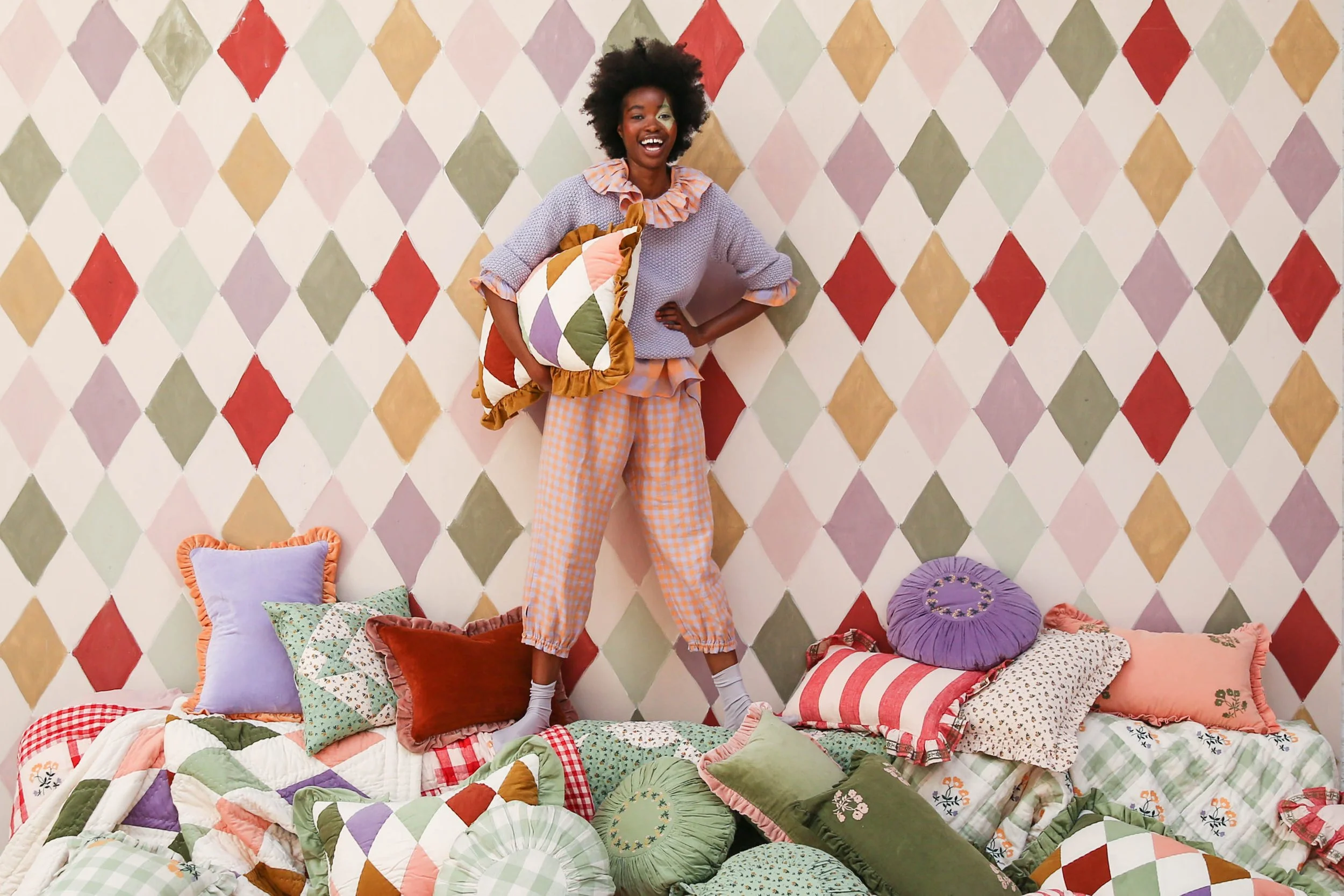 A woman with curly hair standing on a pile of colorful pillows and quilts, smiling in front of a diamond-patterned wall.