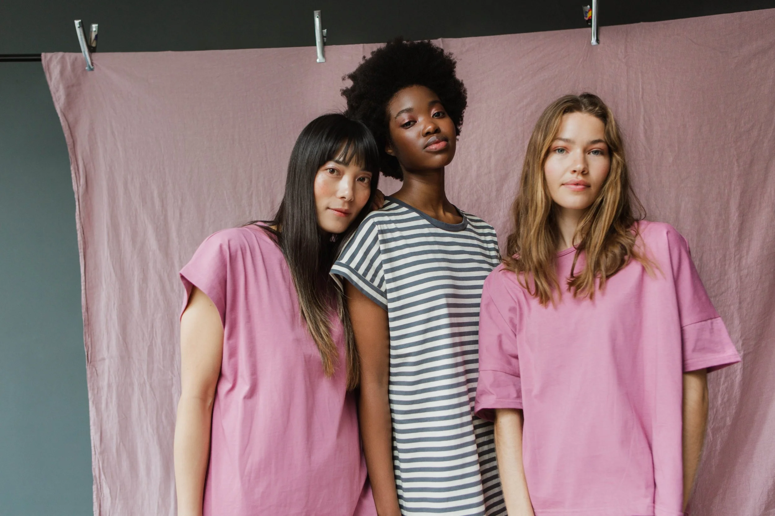 Three women standing in front of a pink backdrop, wearing casual clothing, with diverse appearances and hair types.
