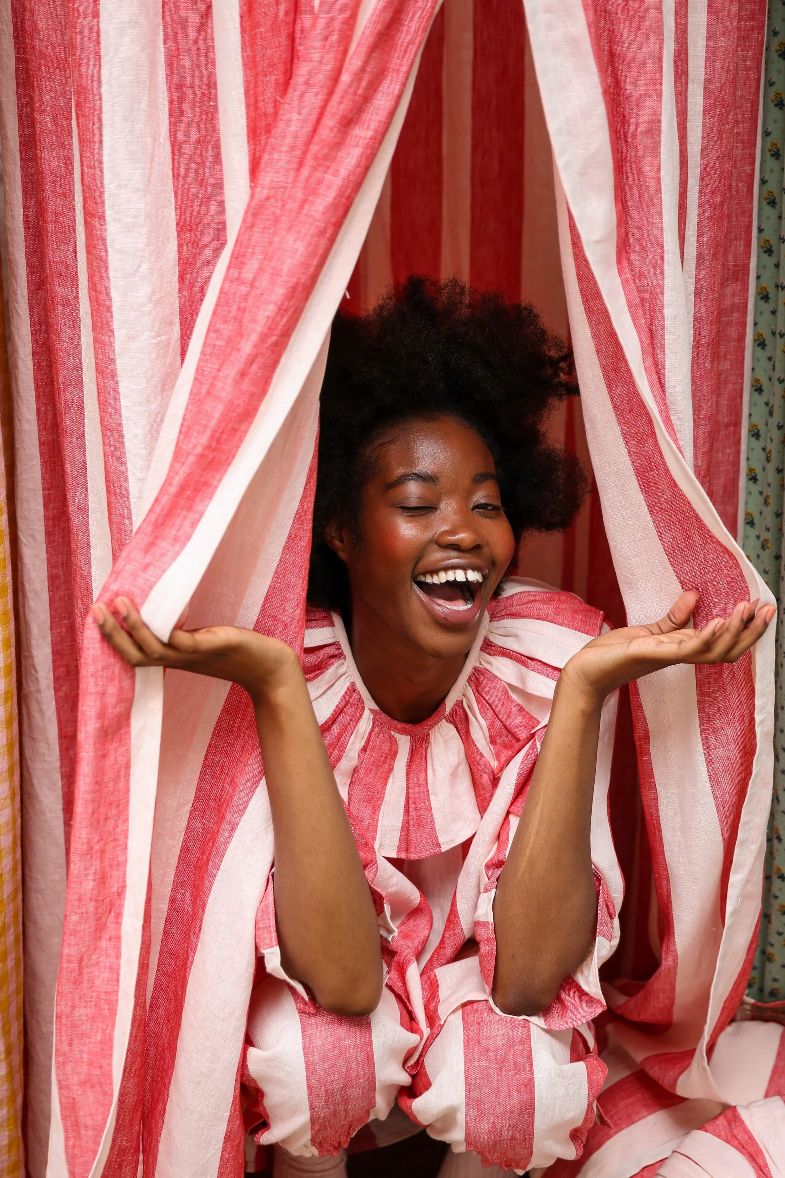 A young woman with curly hair smiling and winking, wearing a red and white striped outfit, leaning out from behind red and white striped curtains.
