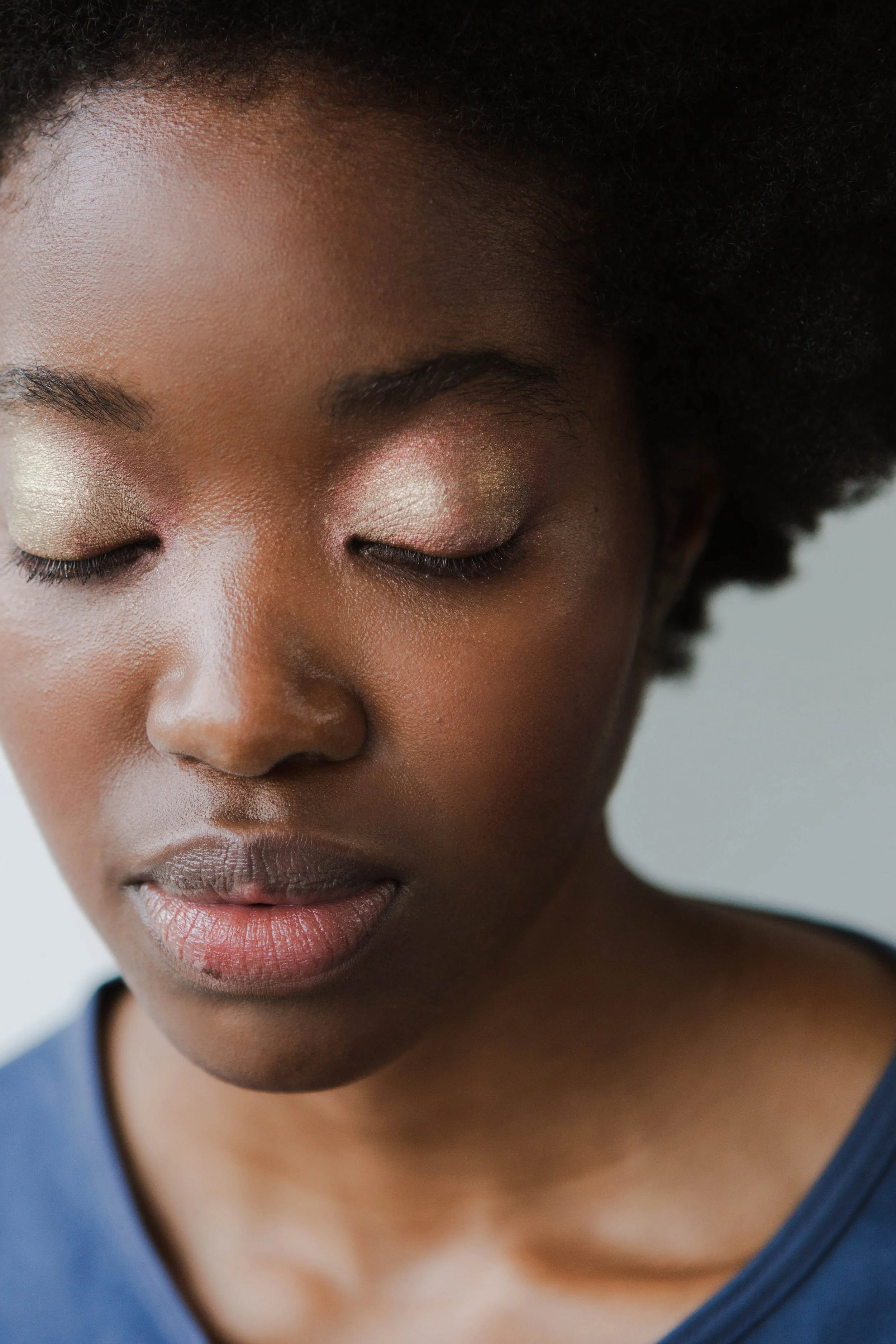 Close-up of a woman with dark skin, eyes closed, wearing gold eyeshadow, with natural curly hair and a blue top.