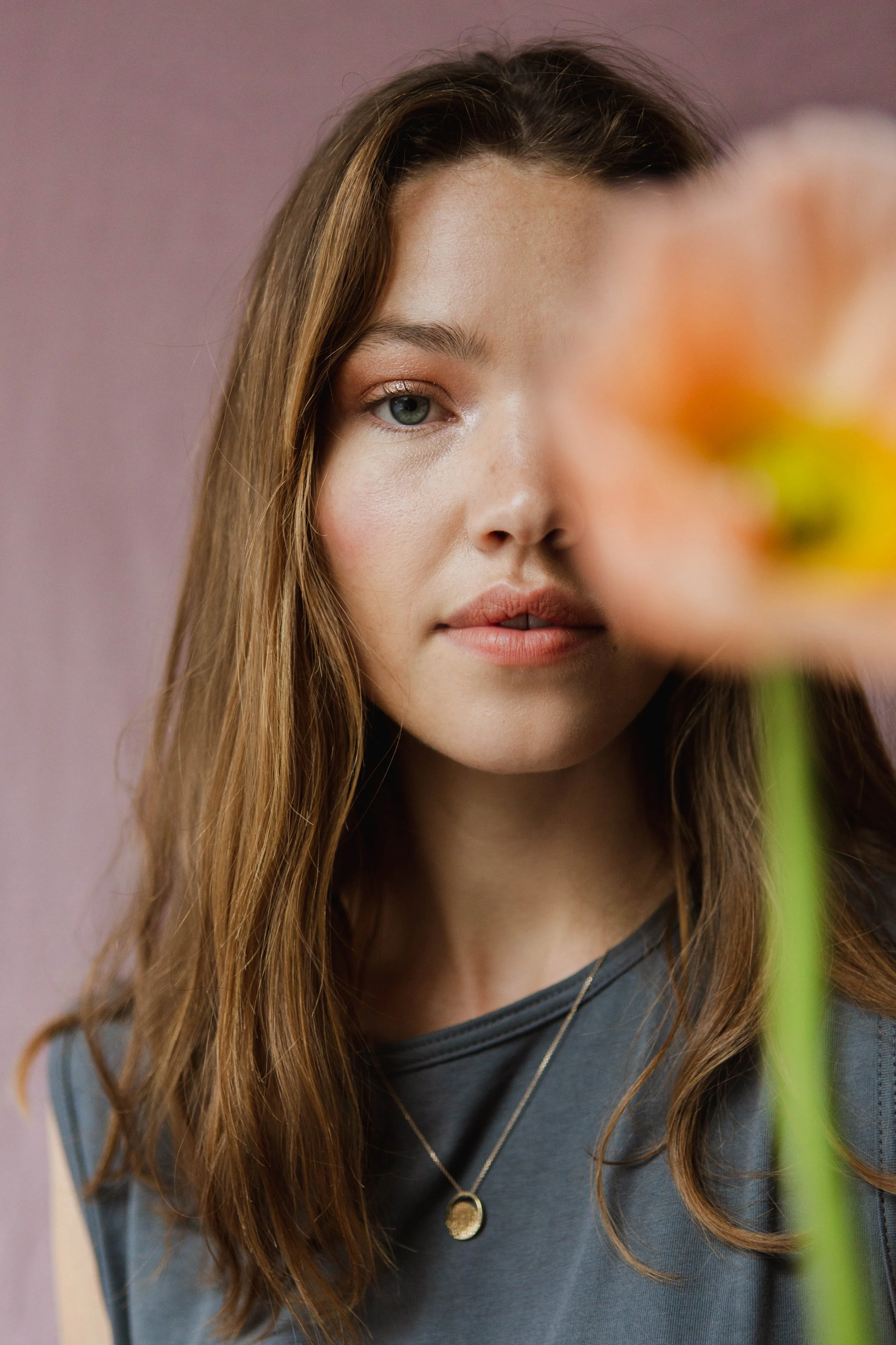 A woman with long brown hair and fair skin looking at the camera behind an out-of-focus orange flower.