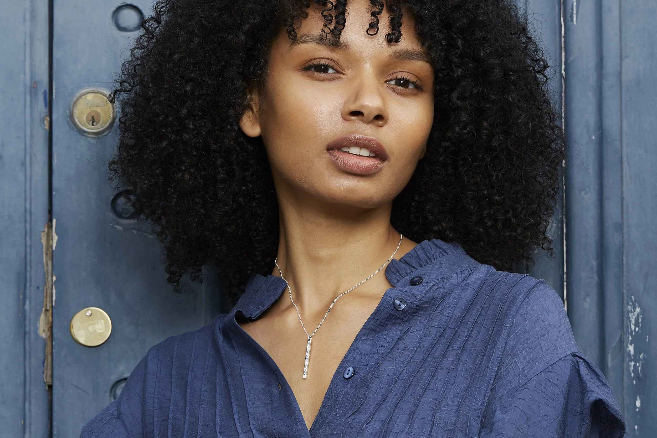 A woman with curly black hair wearing a blue shirt and a silver necklace stands near a blue wooden wall.