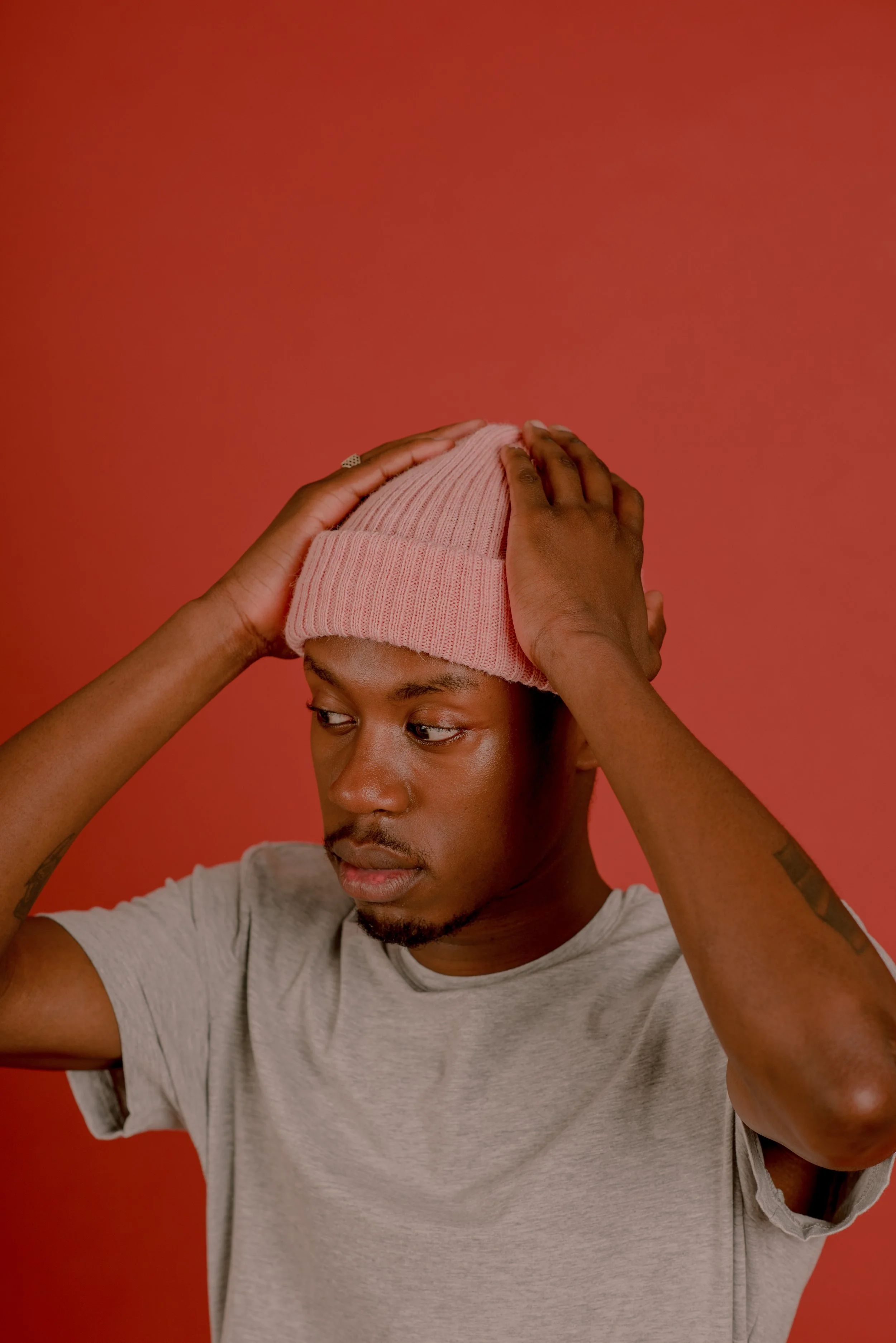 A young man wearing a light gray t-shirt and a pink knit beanie, holding his head with both hands against a reddish background.