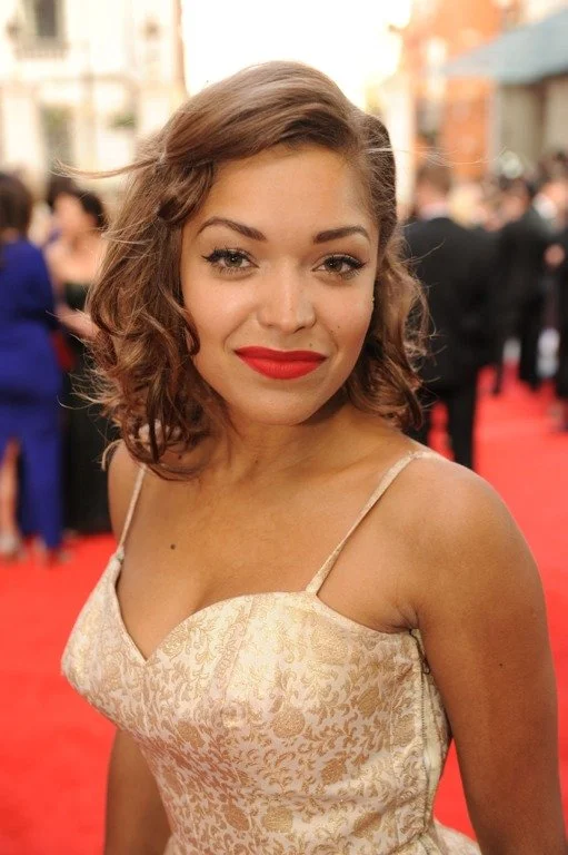 A young woman with shoulder-length wavy brown hair and red lipstick, wearing a beige dress with thin straps, smiling at the camera on a red carpet at an outdoor event.