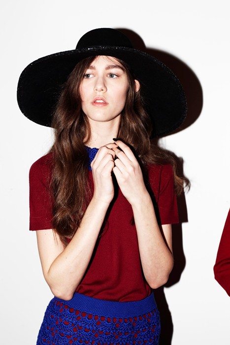 Young woman with long brown hair wearing a large black hat, red top, and blue skirt against a white background.