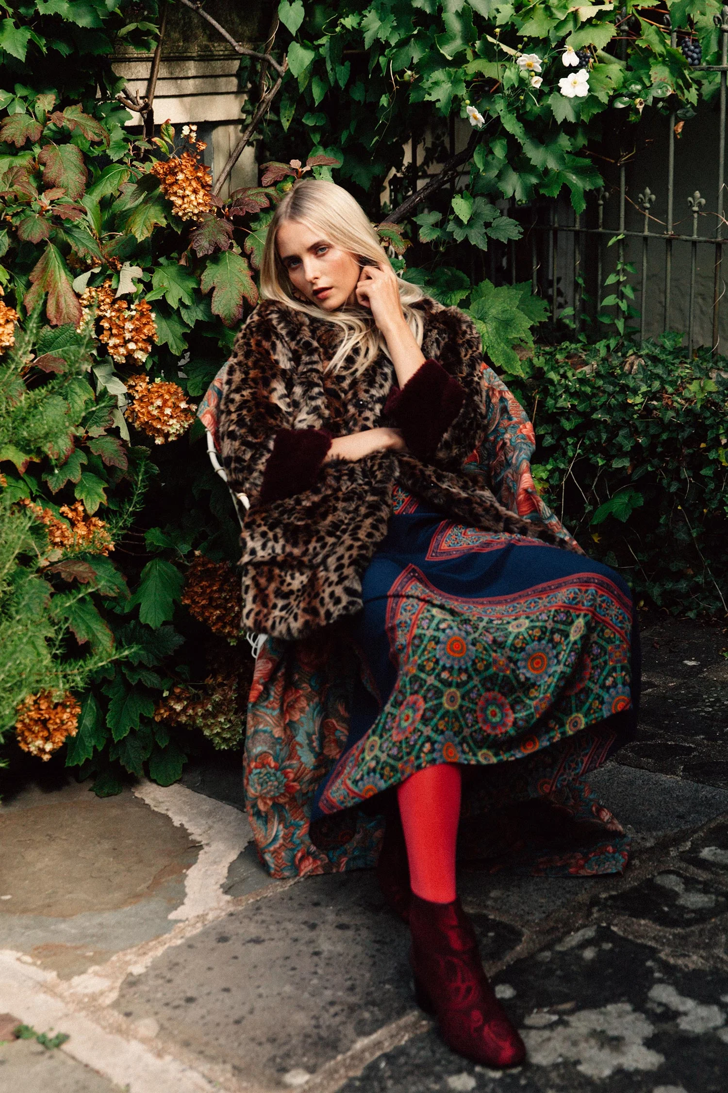 A woman seated outdoors surrounded by lush green and orange foliage, wearing a leopard print fur coat, a colorful patterned long skirt, red tights, and red velvet ankle boots.