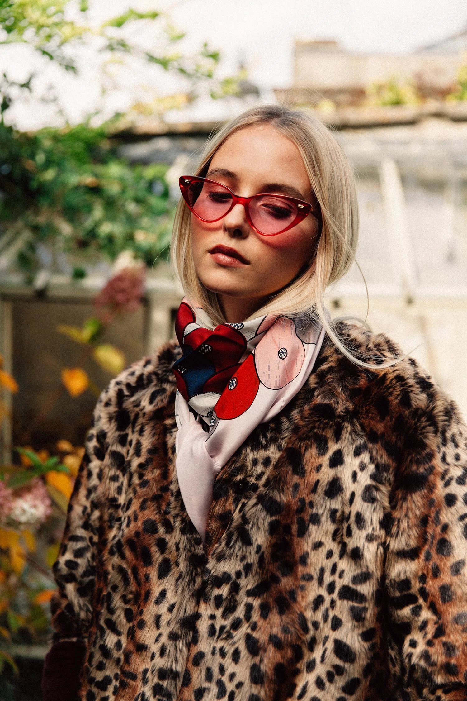 A young woman with blonde hair wearing red sunglasses, a black, white, and red patterned scarf, a leopard-print coat, and standing outdoors among greenery.