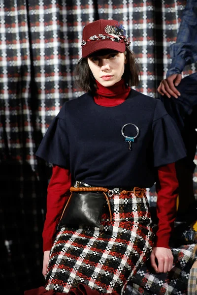Model wearing a red cap, navy top with ruffle sleeves, red turtleneck, plaid skirt, and a black pouch with a gold chain at a fashion show.