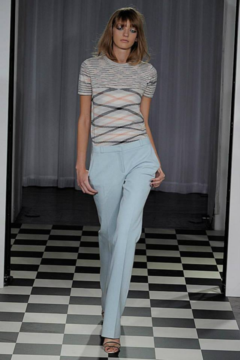 Woman with shoulder-length hair and bangs wearing a striped shirt with a diamond pattern and light-colored pants walking on a black and white checkered floor.