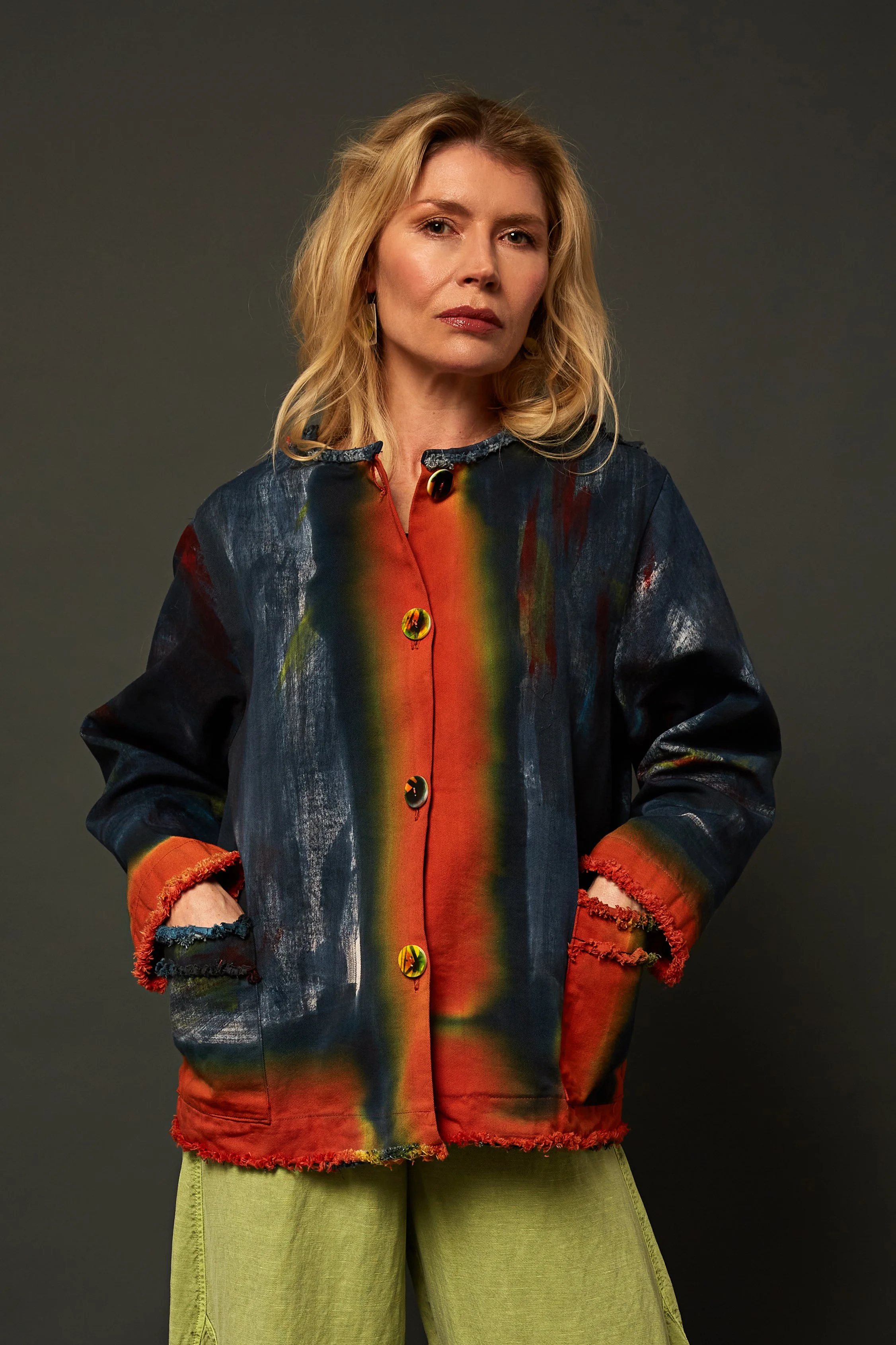 A woman with blonde hair wearing a colorful, patterned jacket with frayed edges and light green pants standing against a dark gray background.
