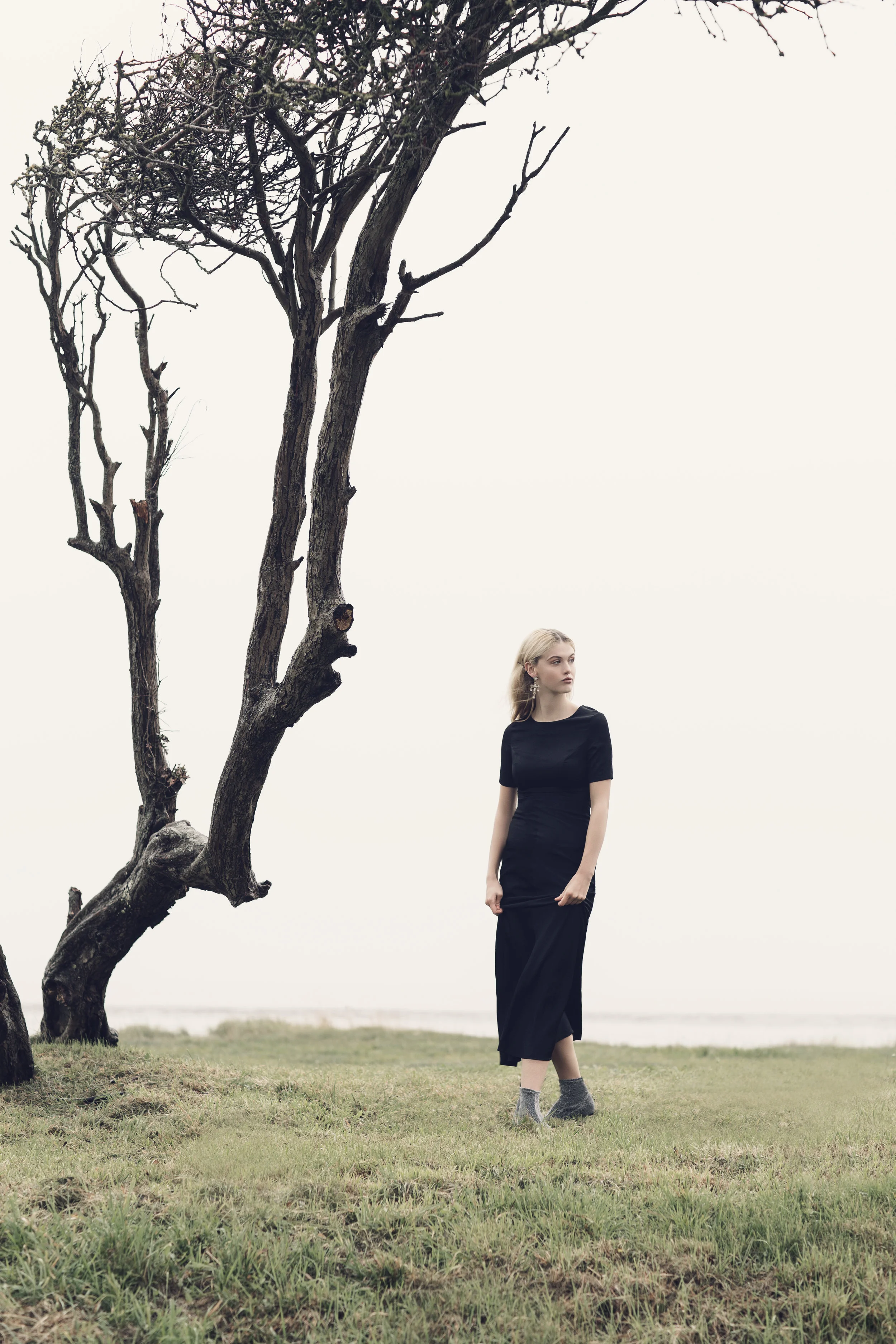A young woman in a black dress and gray boots stands on a grassy field next to a leafless, twisted tree on a cloudy day.