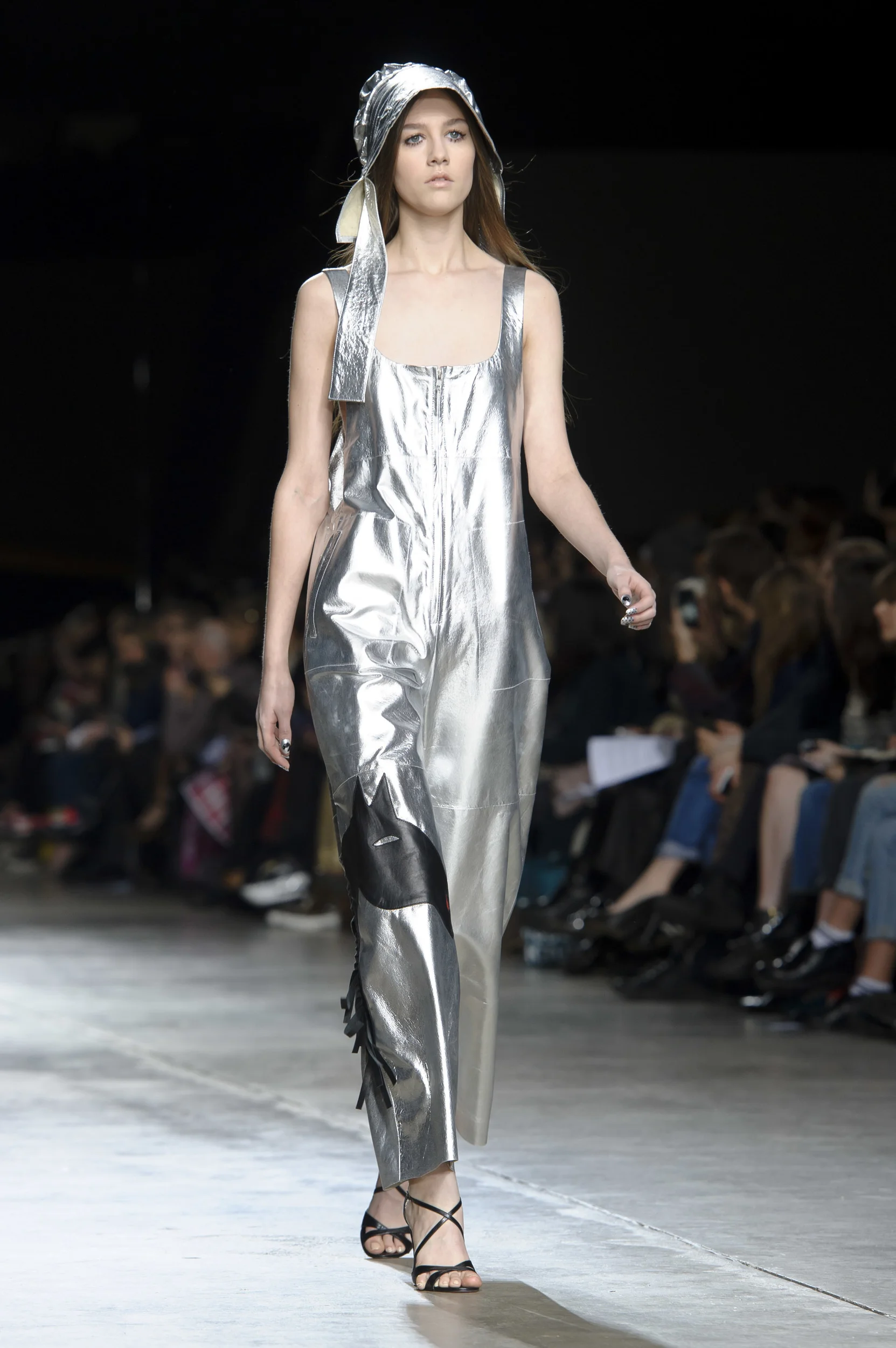 A female model walking down a runway during a fashion show, wearing a shiny, metallic silver jumpsuit and a matching silver headscarf, with black open-toe high heels.