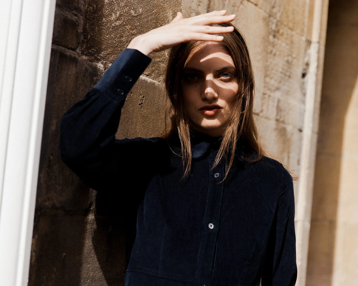 A woman in a black shirt leaning against a textured stone wall, shielding her eyes from sunlight with her hand.