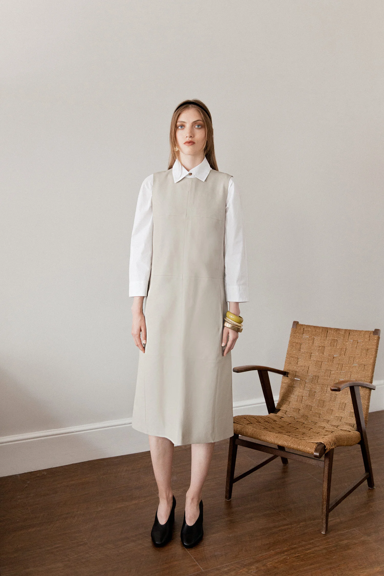 A woman with long light brown hair and blue eyes standing indoors, wearing a white blouse under a beige sleeveless dress that extends below her knees, black high heels, and gold bangles on her left arm, next to a mid-century style wooden chair with w
