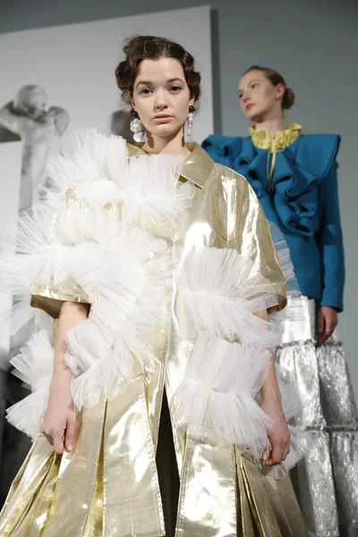 Two women wearing elaborate high-fashion outfits, one in a gold and white dress with tulle accents and large earrings, the other in a striking blue jacket with exaggerated shoulders, standing in an art gallery setting.