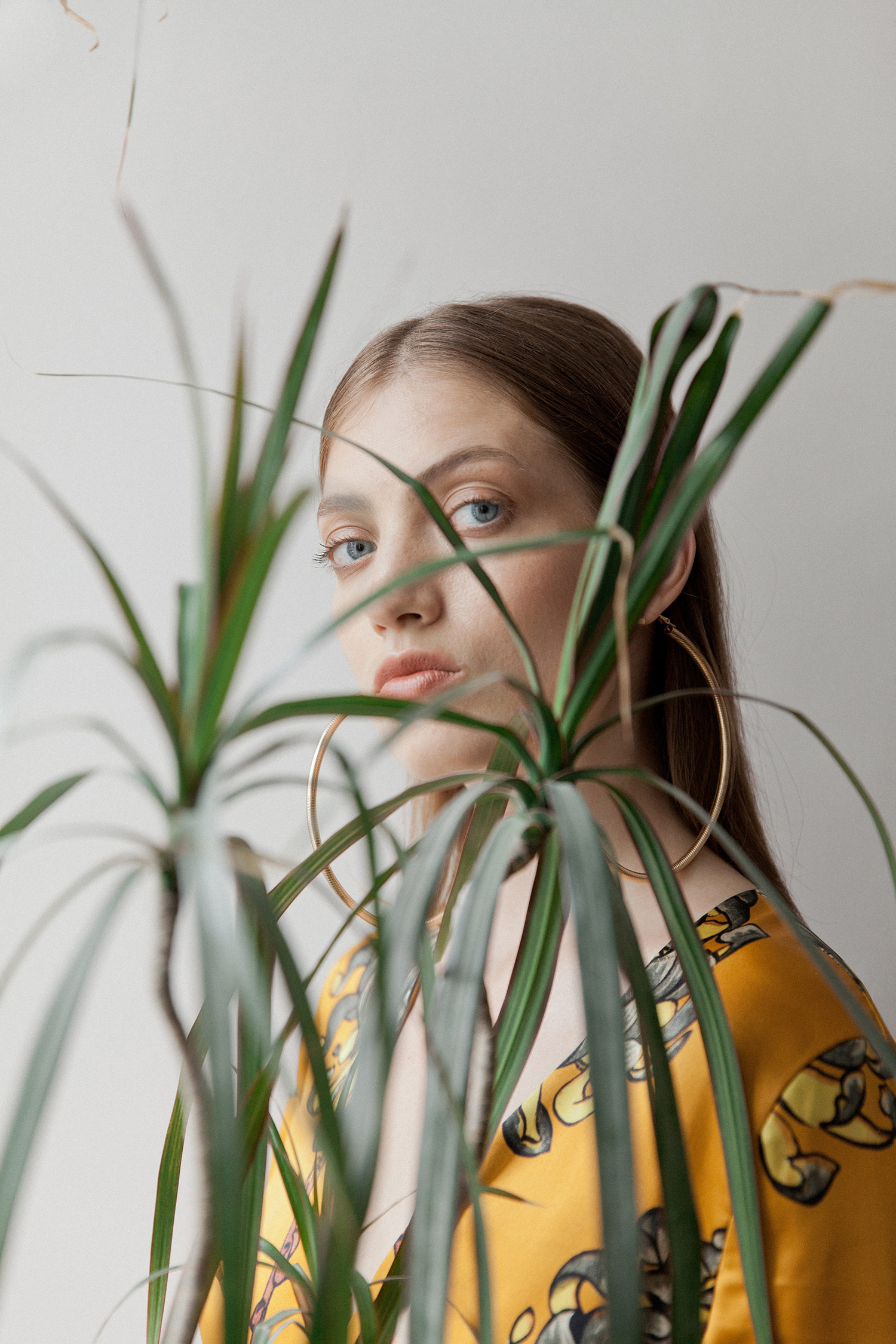 A young woman with brown hair, blue eyes, wearing large gold hoop earrings and a yellow patterned top, stands behind green leafy plants, partially obscuring her face.