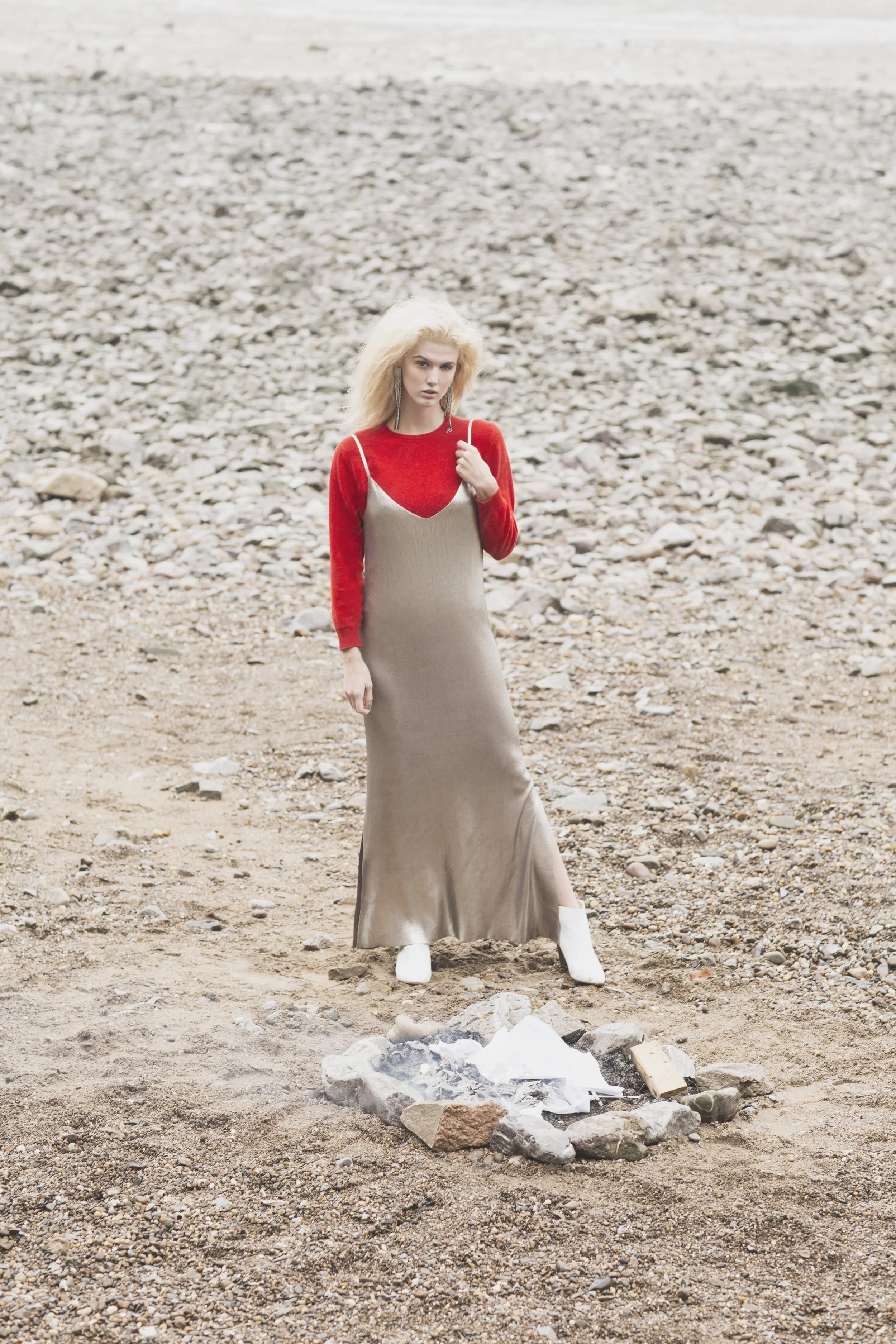 Woman in a beige dress and red sweater standing on a rocky beach near a small fire pit.