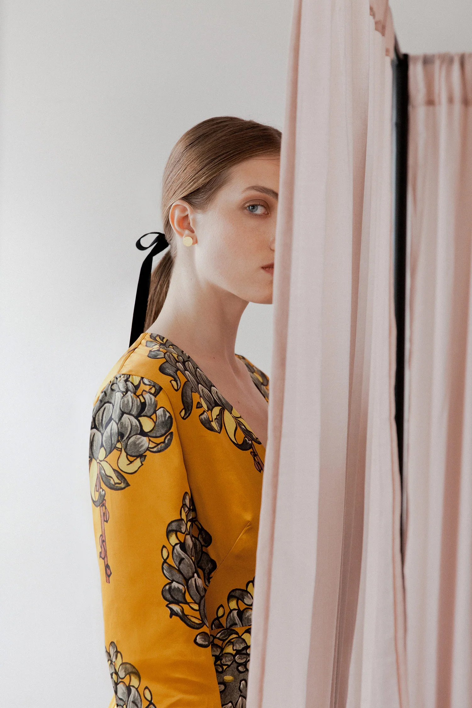 A woman in a yellow floral dress trying on clothes at a fitting room, partially hidden behind a curtain.