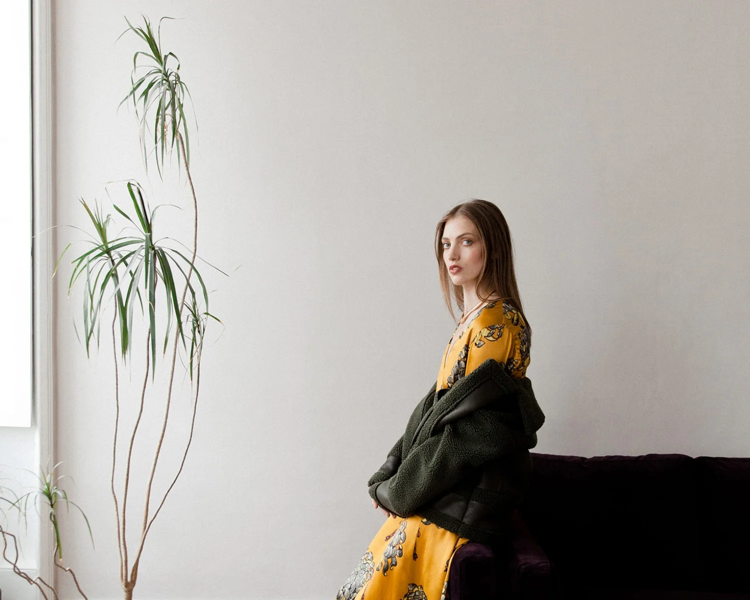 A woman with brown hair wearing a yellow floral dress and a green jacket, sitting on a purple sofa beside a tall houseplant, in a room with plain white walls and a window.