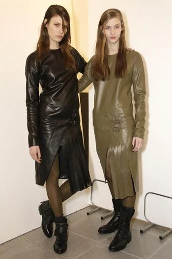 Two young women wearing leather outfits, one in black and the other in olive green, standing indoors against a plain wall.