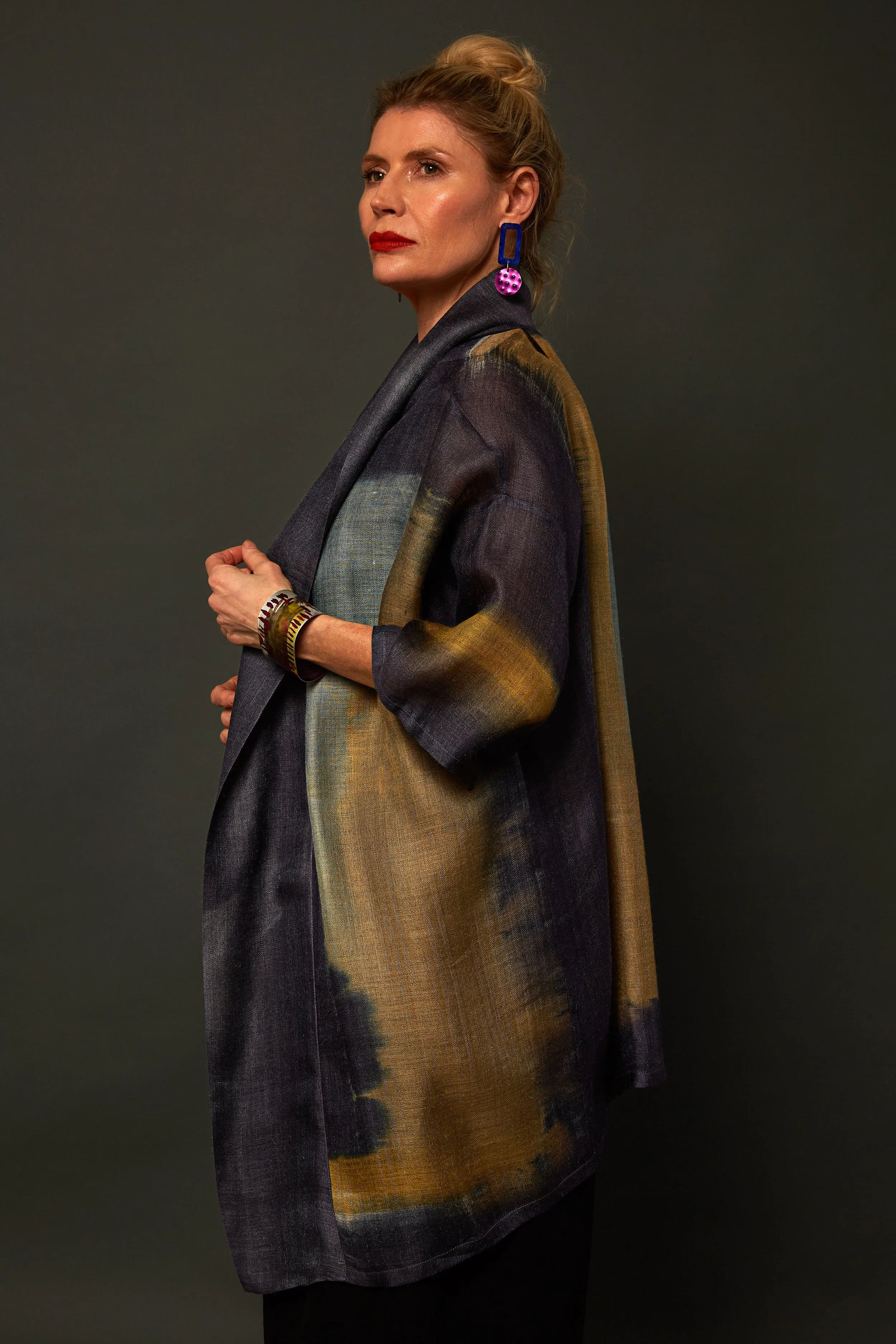 A woman with red hair styled in an updo, wearing bold red lipstick and colorful earrings, poses against a dark background. She is dressed in a multicolored, loose-fitting, abstract-patterned jacket and accessorized with bracelets.