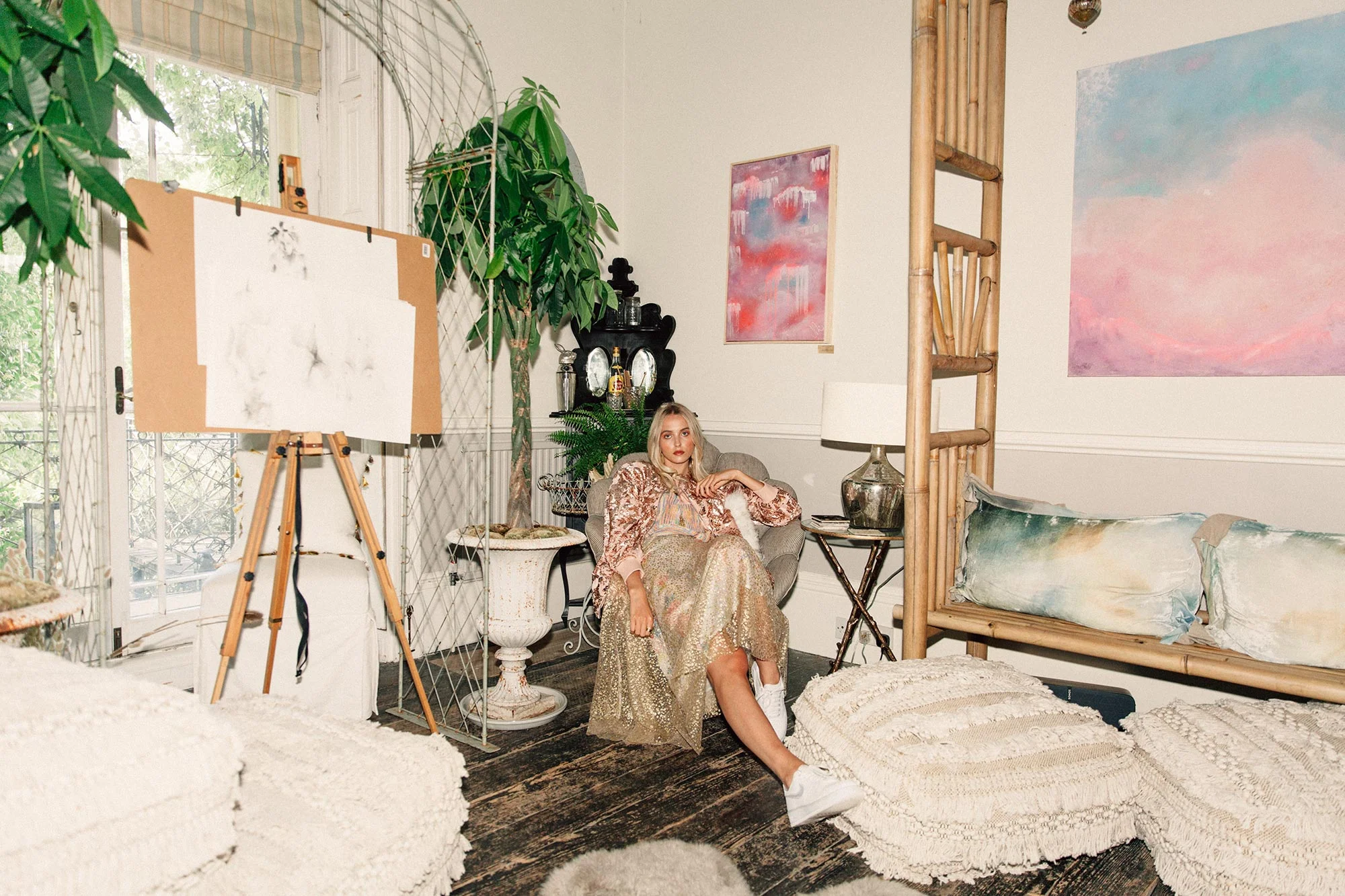 A woman with blonde hair sitting in an eclectic living room with large paintings on the wall, vintage furniture, plants, and a canvas on an easel.