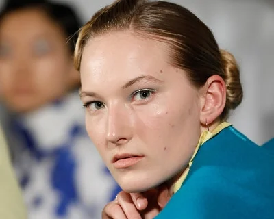 Close-up of a woman with reddish hair in a bun and blue eyes, wearing a blue shirt, with a serious expression. Blurred person in background.