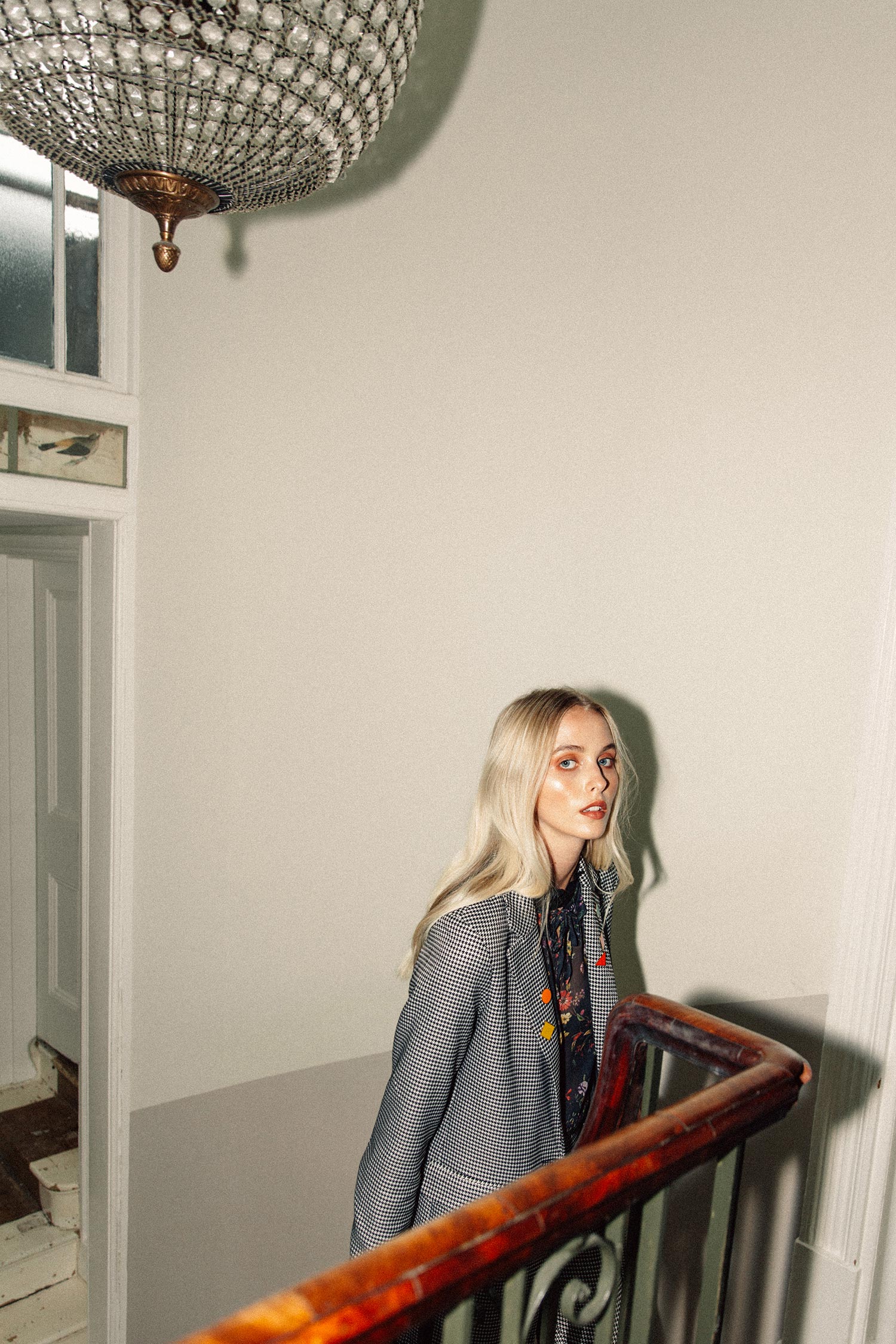 A woman with blonde hair and makeup standing on a staircase in a house, wearing a checkered jacket over a patterned shirt, looking at the camera.