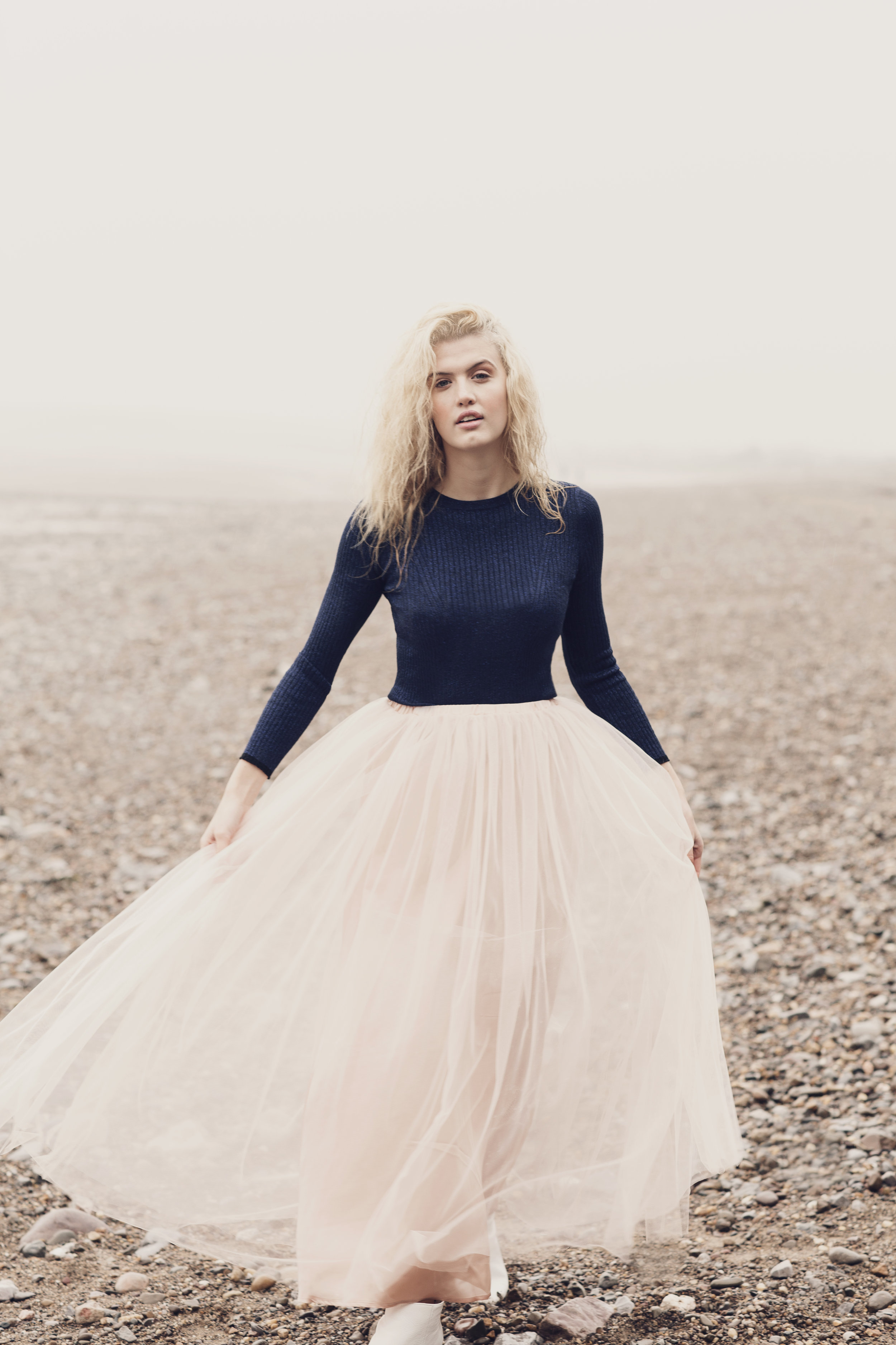 A woman with curly blonde hair standing on a pebble beach, wearing a navy blue long sleeve top and a long, light-colored tulle skirt.