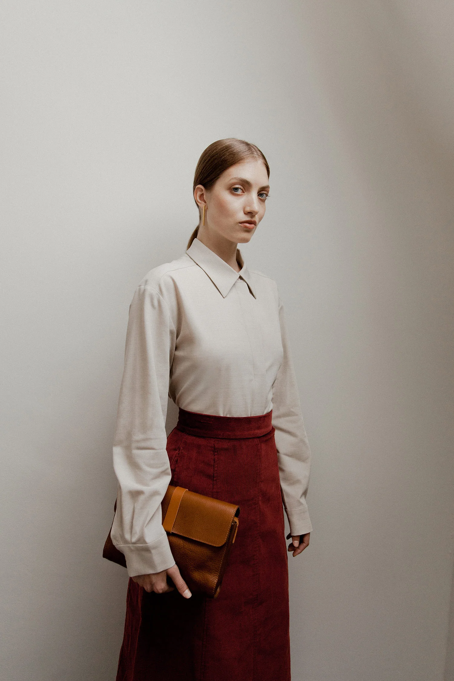 A woman stands against a plain wall, wearing a beige collared shirt and a long red skirt, holding a brown clutch purse.