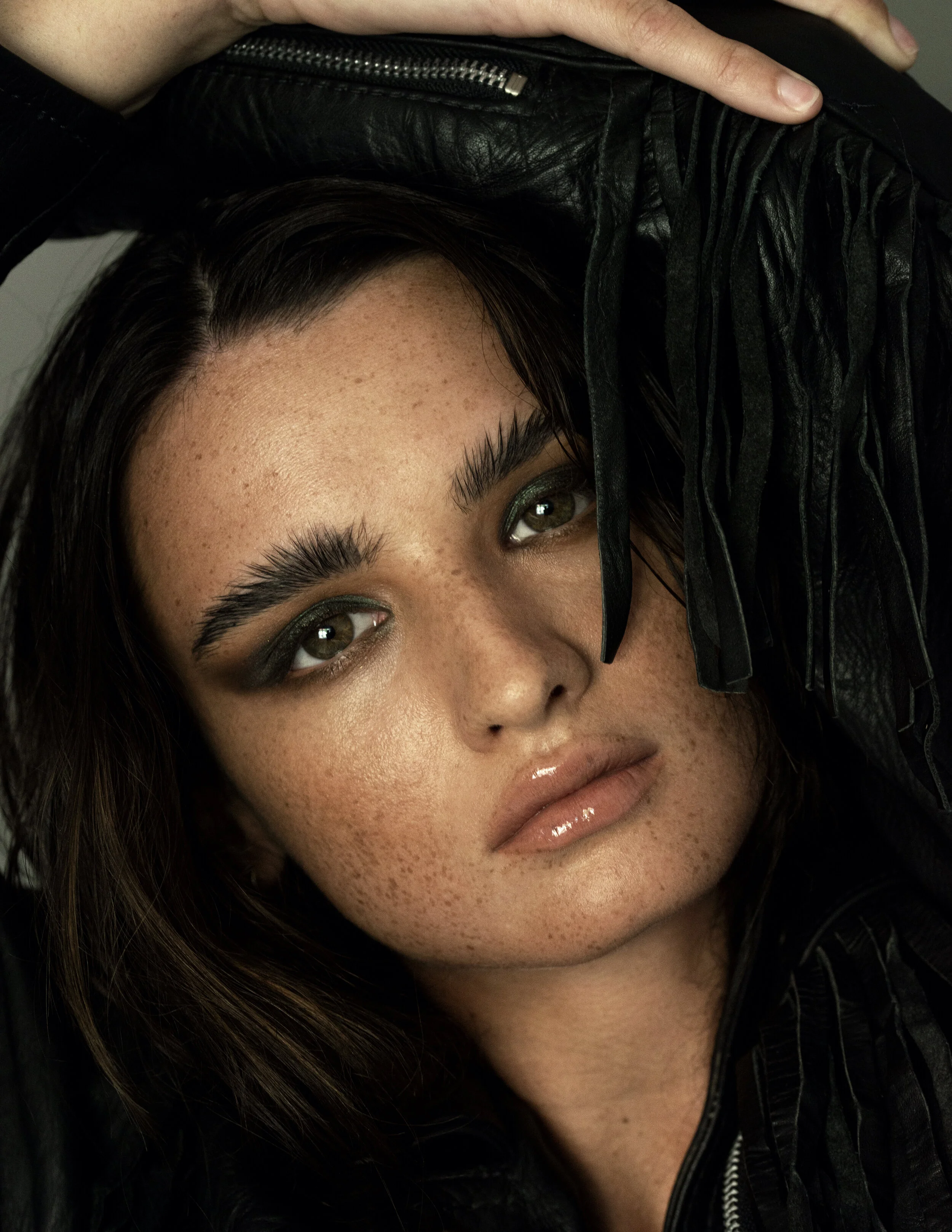 Close-up portrait of a woman with dark hair, blue eyes, and freckles, wearing shiny makeup and a black leather jacket, with her hand resting on her head.