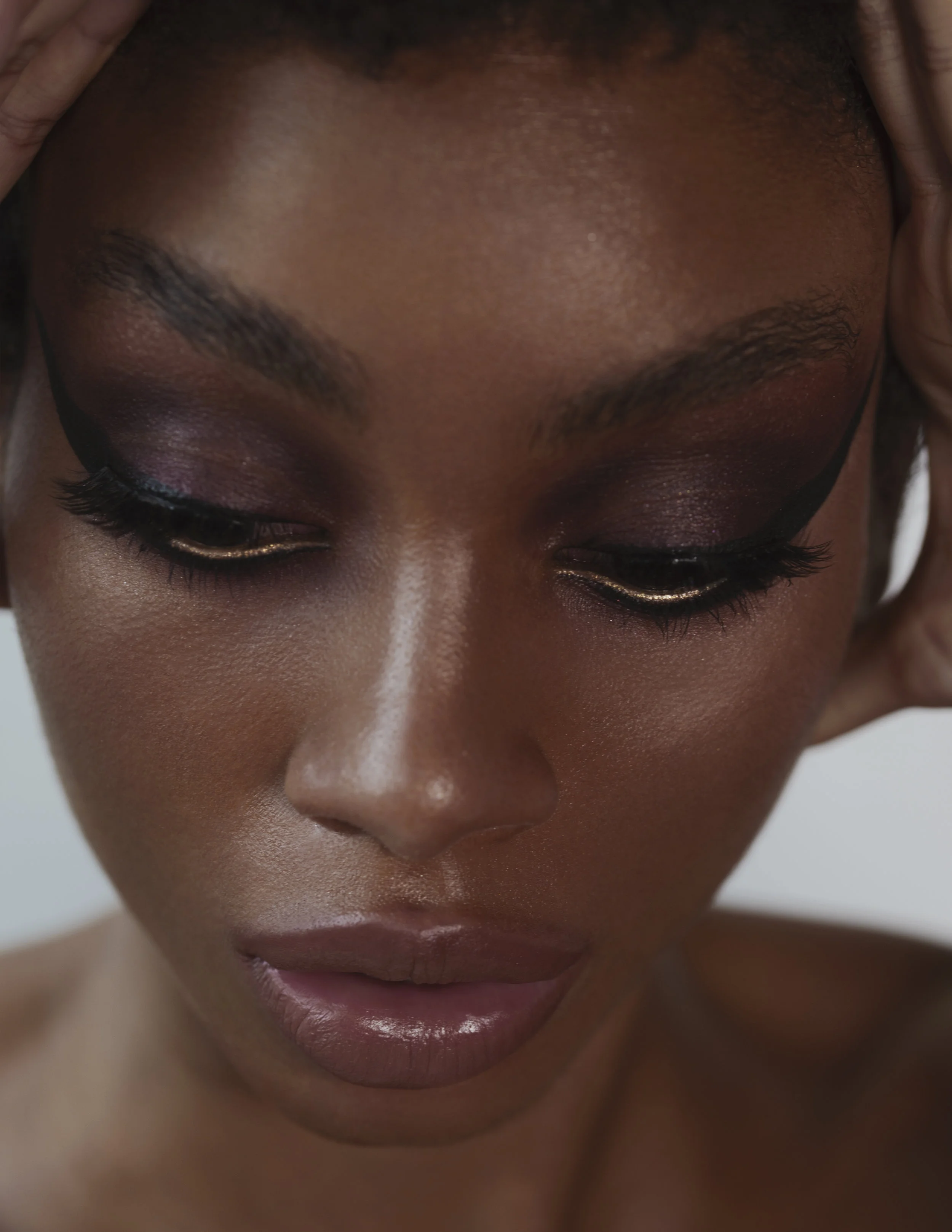 Close-up of a woman's face with dark skin, wearing purple eyeshadow and gold eyeliner, with her eyes closed and hands on her temples.