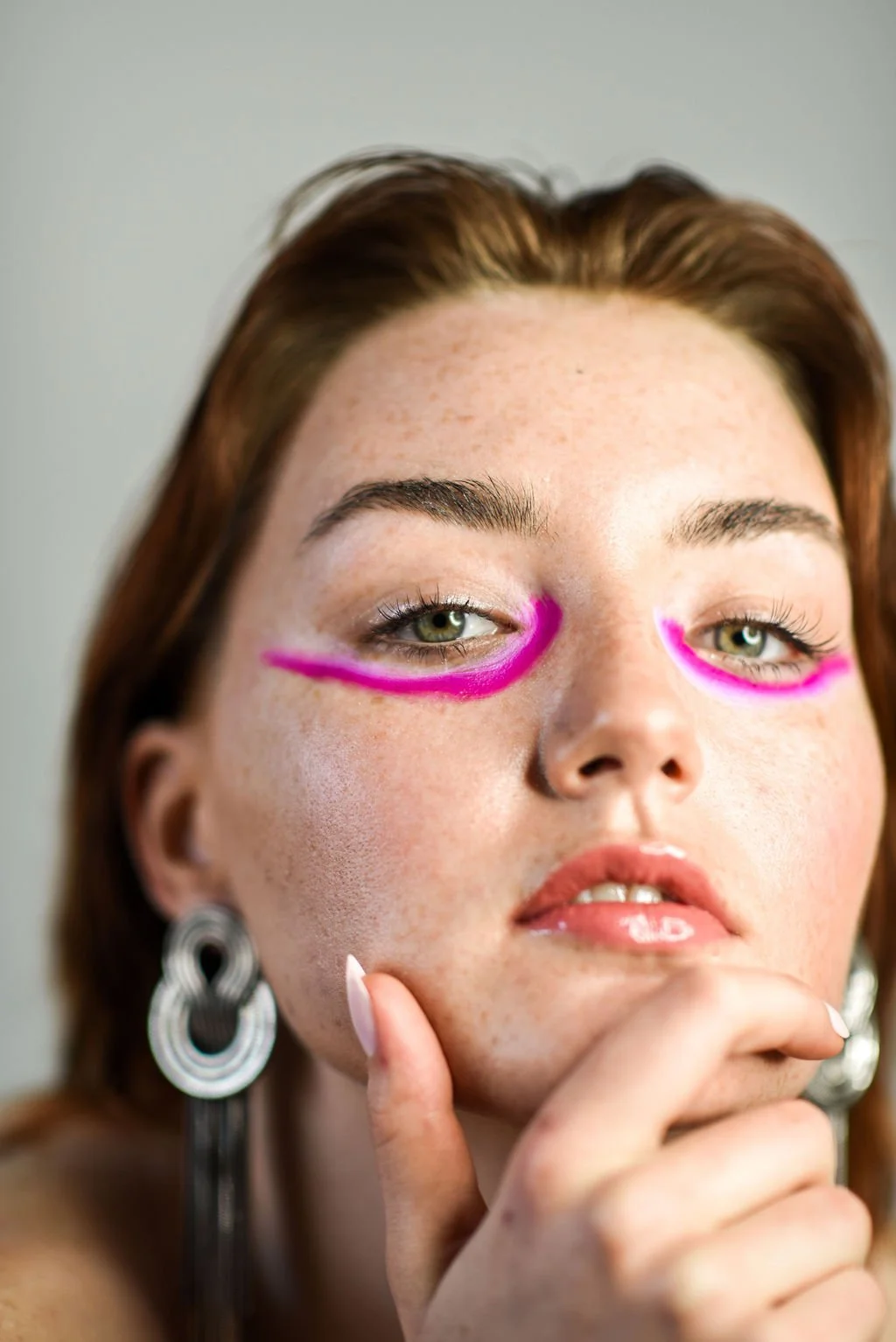 A close-up of a woman's face with neon pink makeup lines under her eyes, wearing large chain earrings, and touching her chin with her hand.