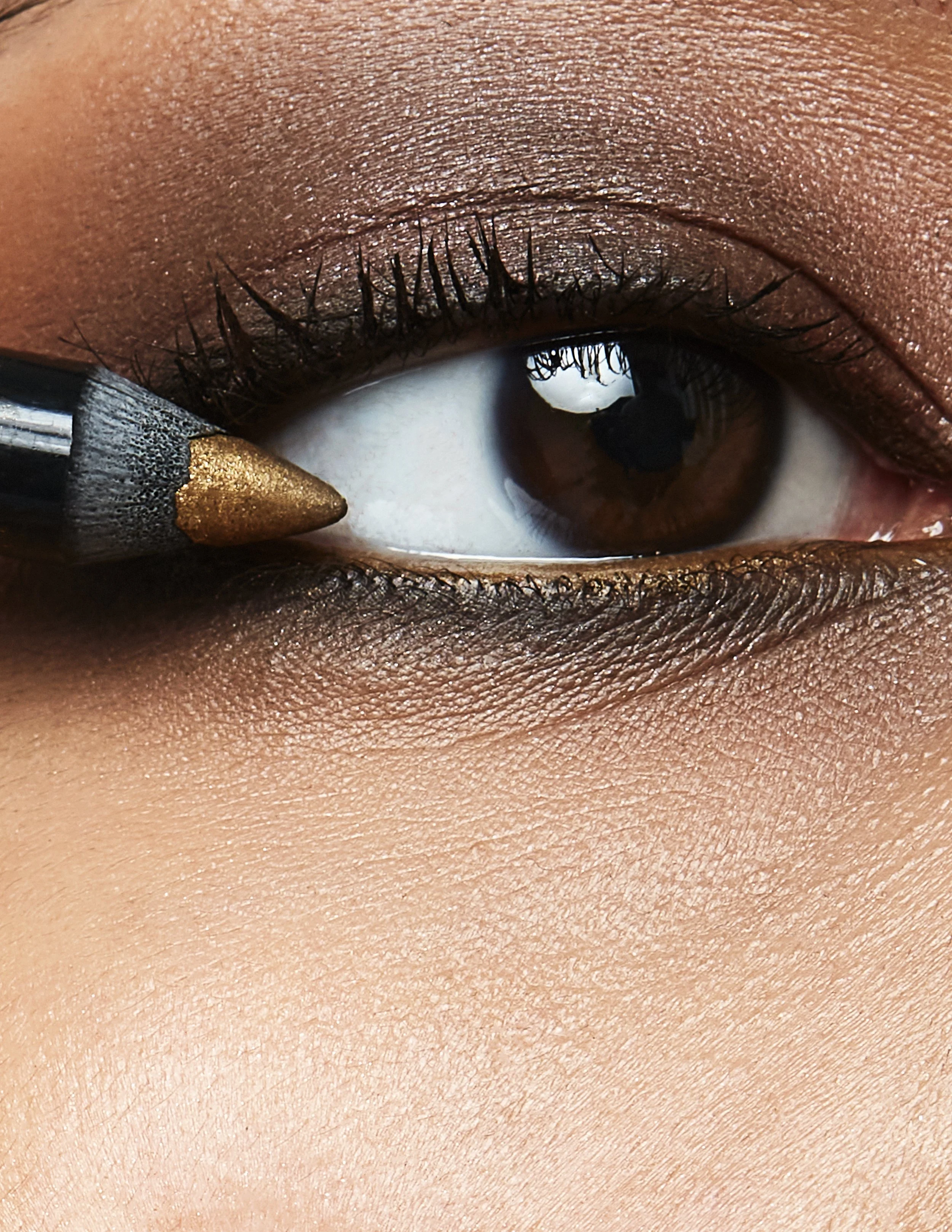 Close-up of a brown eye with makeup, applying gold eyeshadow with a pencil