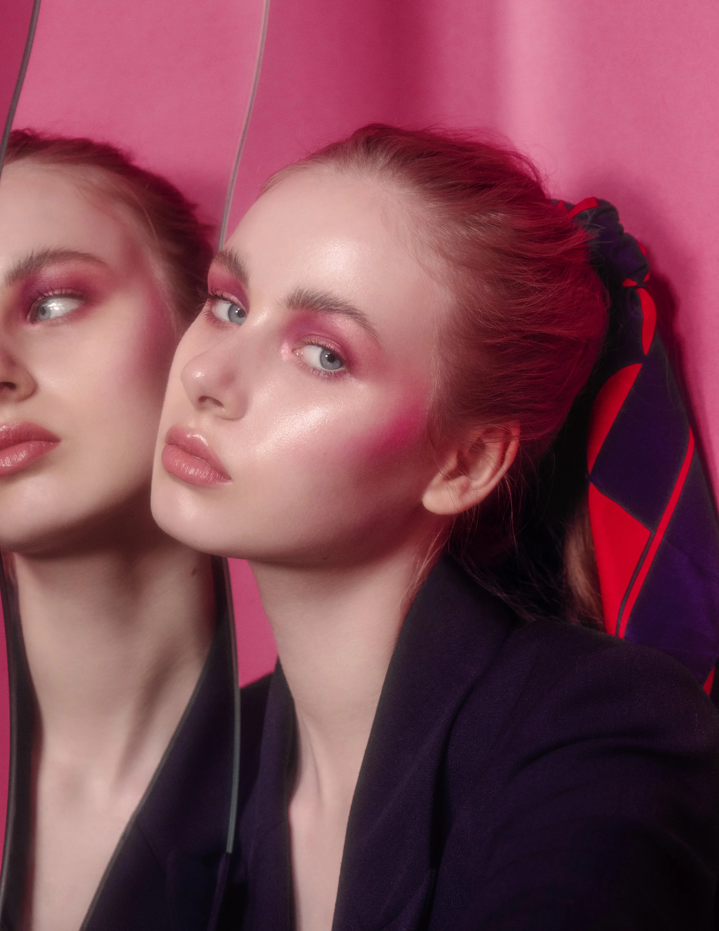 A woman with blonde hair tied in a ponytail, wearing makeup with pink eye shadow and lipstick, gazes at her reflection in a mirror against a pink wall.