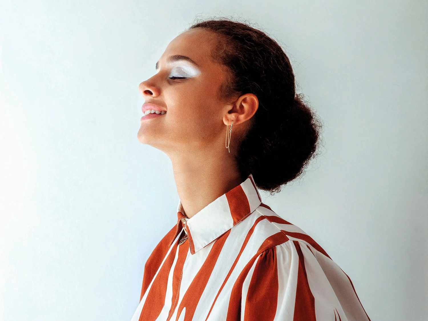 Side profile of a smiling woman with closed eyes, wearing white eyeshadow, a striped shirt with orange and white colors, and a long earrings, standing against a light-colored background.