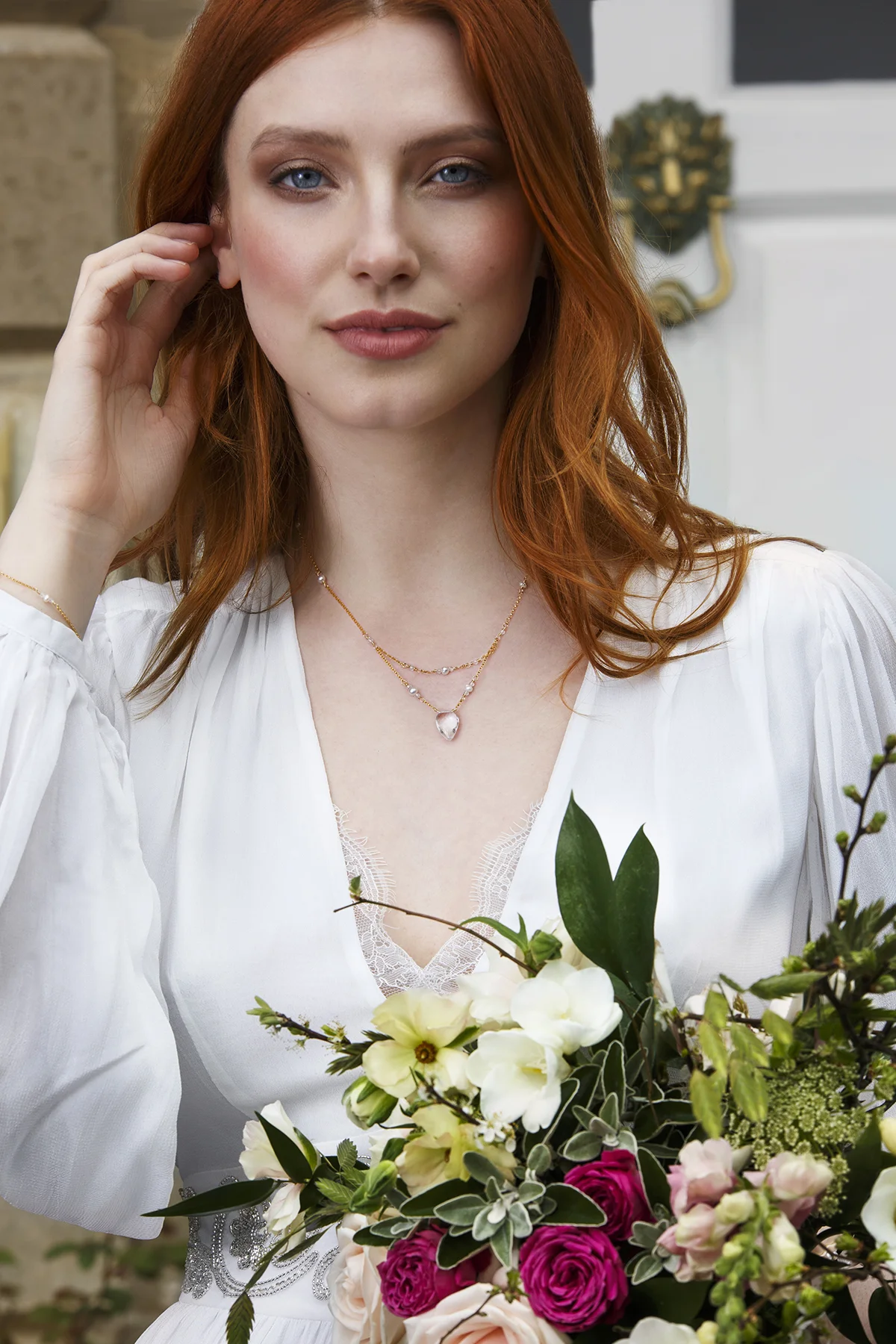 A woman with red hair, blue eyes, and light skin smiling, wearing a white dress and layered necklaces, holding a bouquet of pink, white, and green flowers.
