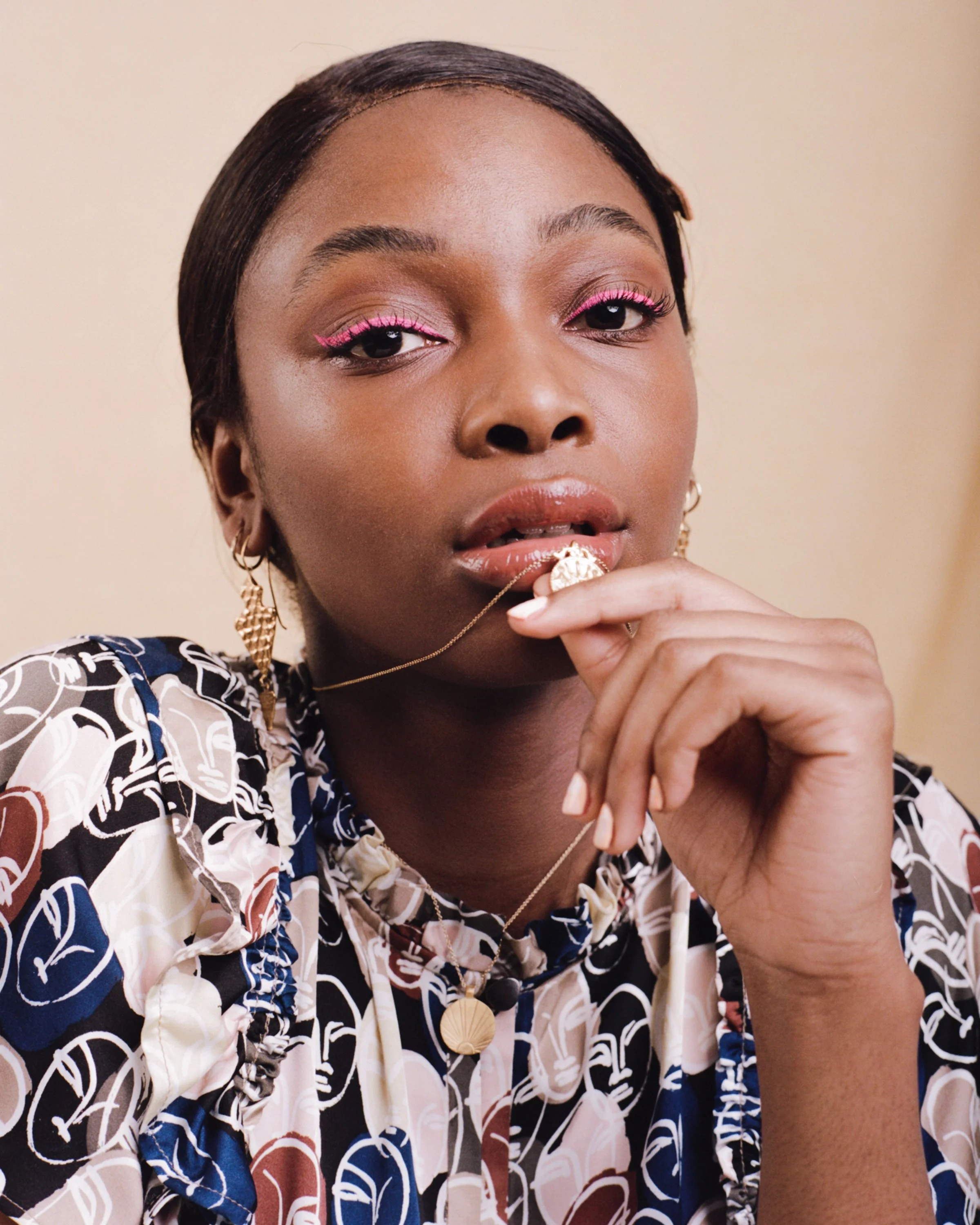 A young woman with short dark hair and gold earrings gazes at the camera, wearing pink eyeliner and patterned clothing, holding a gold necklace with a shell pendant near her lips.