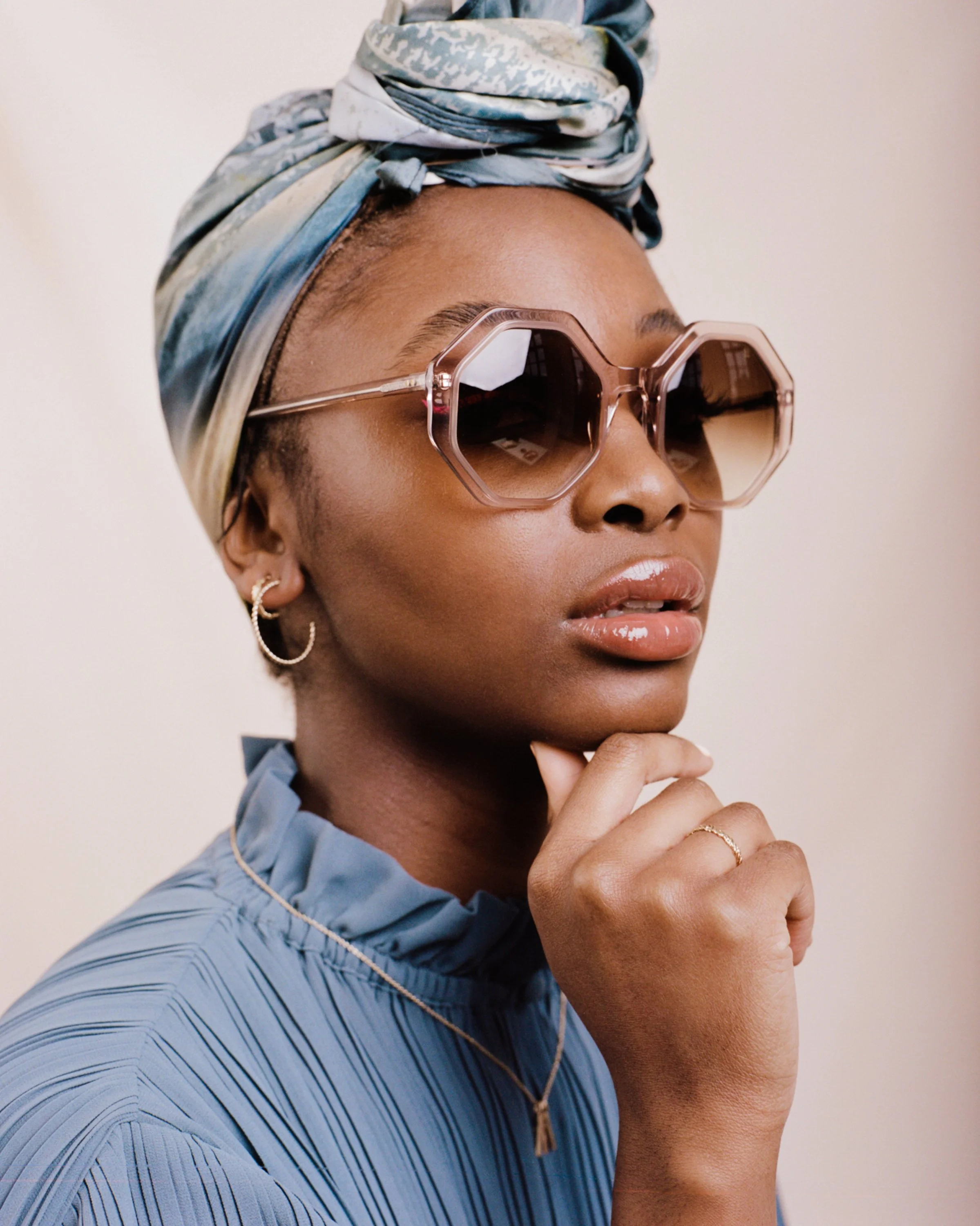 A woman wearing large pink hexagonal sunglasses, a blue headwrap, a light blue pleated blouse, and jewelry including earrings, a necklace, and a ring.