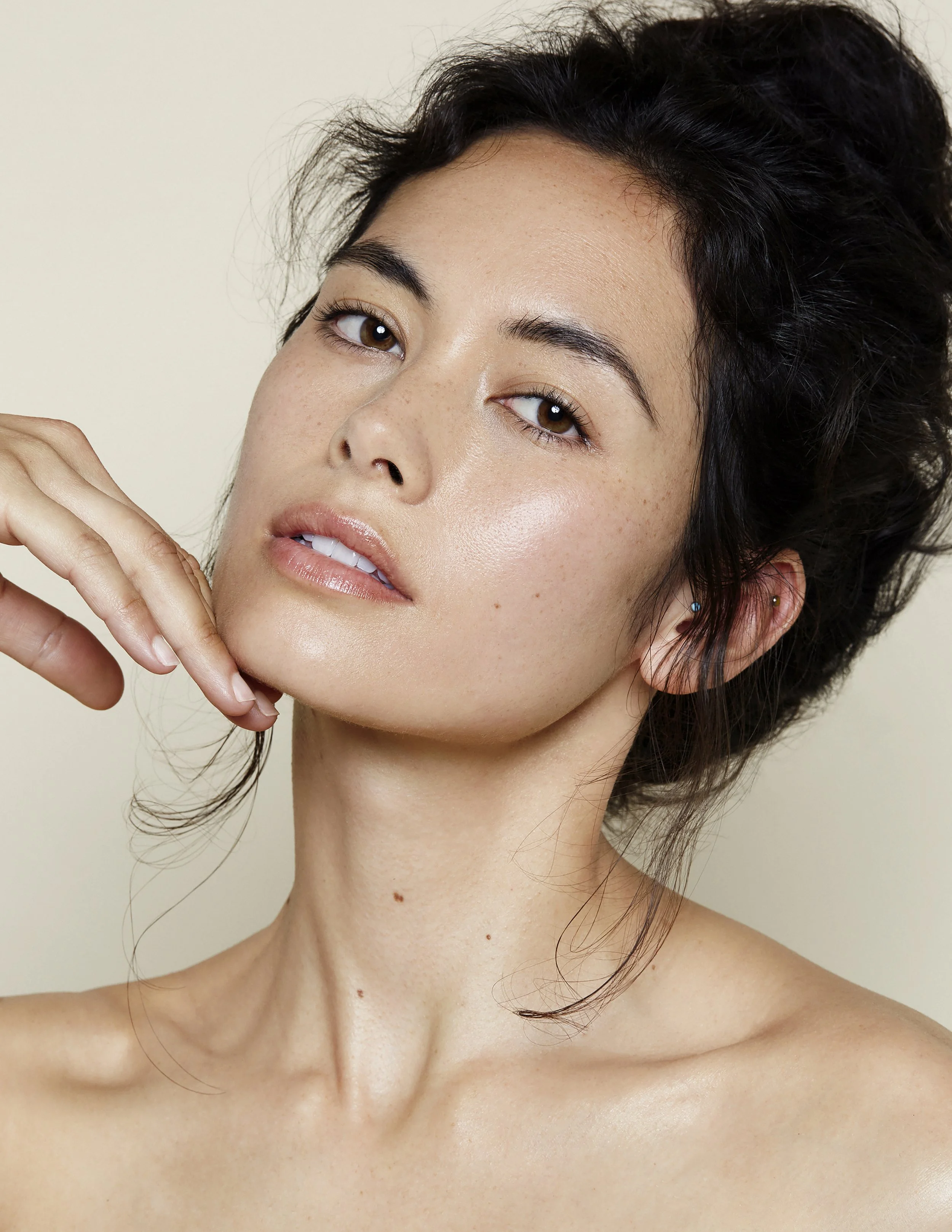 Close-up of a woman with fair skin, brown eyes, dark wavy hair, and natural makeup, touching her chin with her hand against a neutral background.
